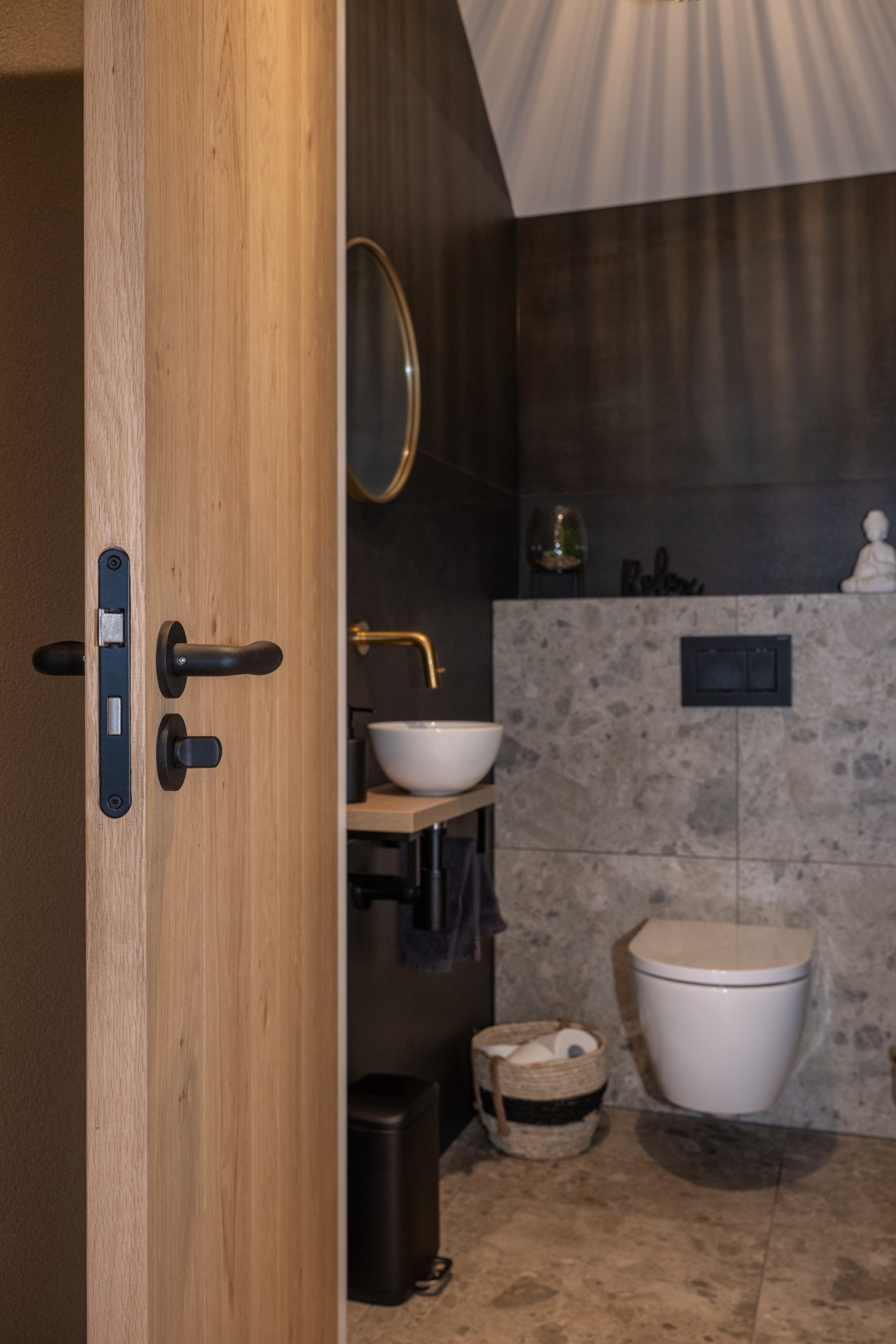 Interieur van een modern toilet met een eikenhouten binnendeur, natuurstenen waskom en goudkleurige kraan.