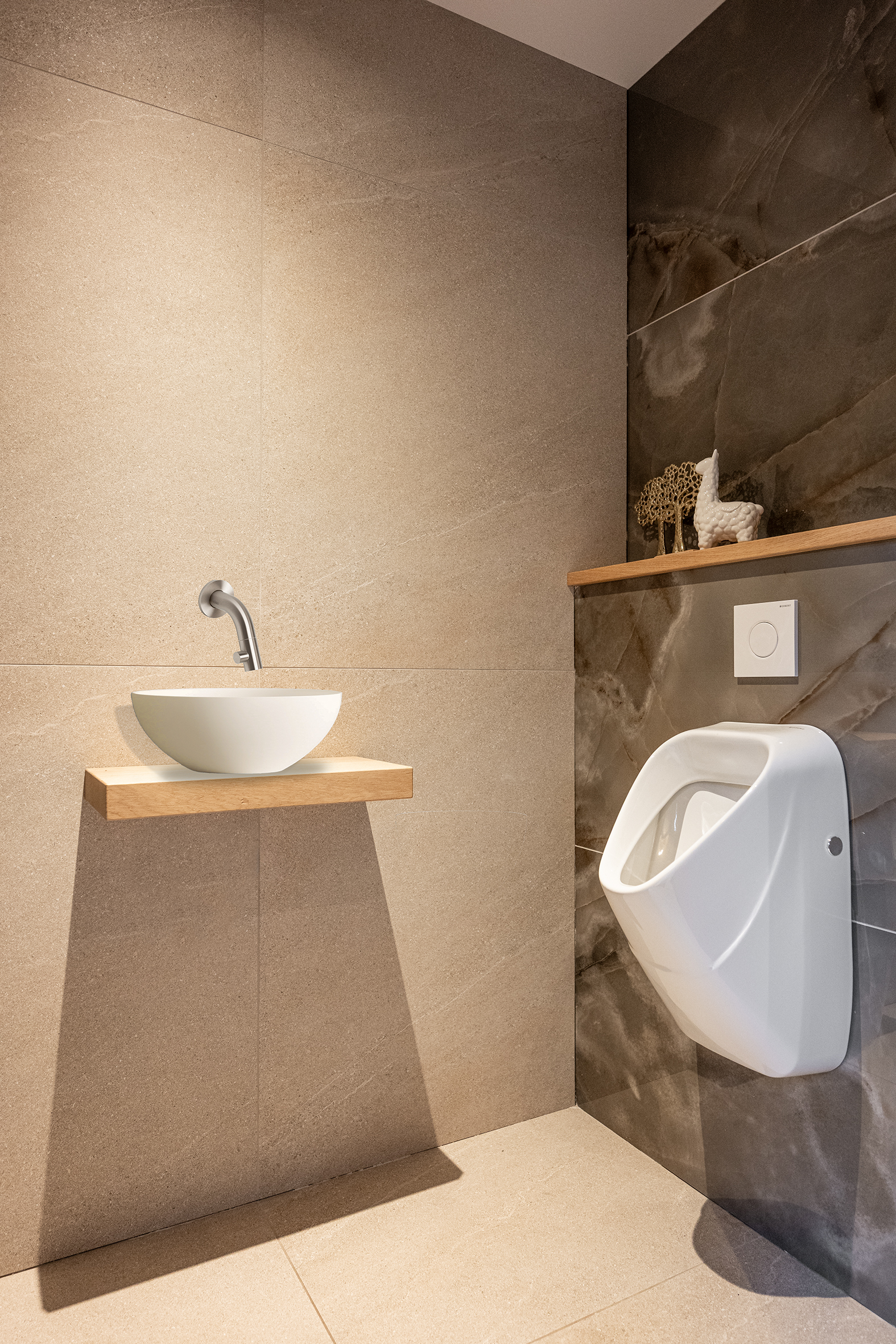 Modern toiletinterieur met een wit urinoir, een witte waskom op een eikenhouten planchet en grote beige wandtegels.