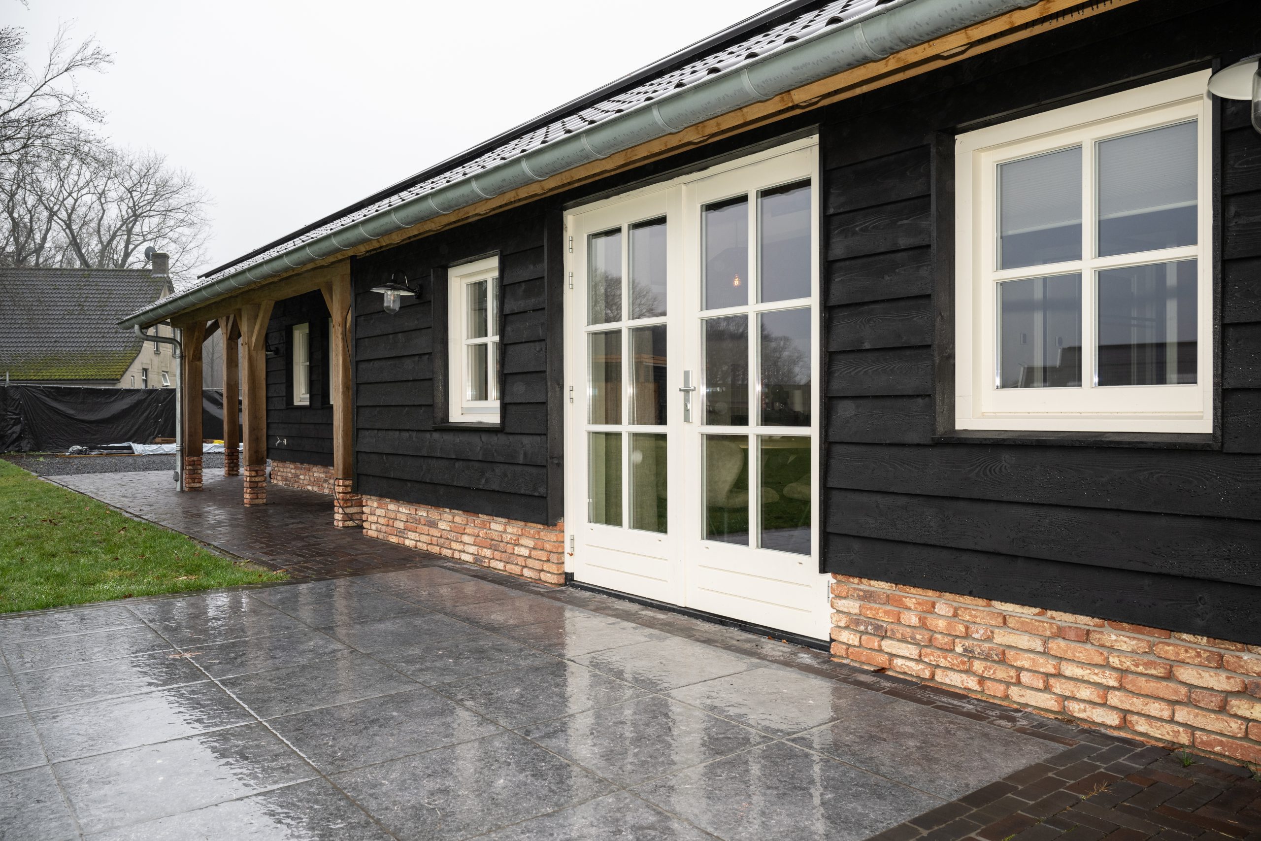 Exterieur van een moderne barn-woning met zwarte houten gevelbekleding, witte kozijnen en een robuuste eikenhouten overkapping.