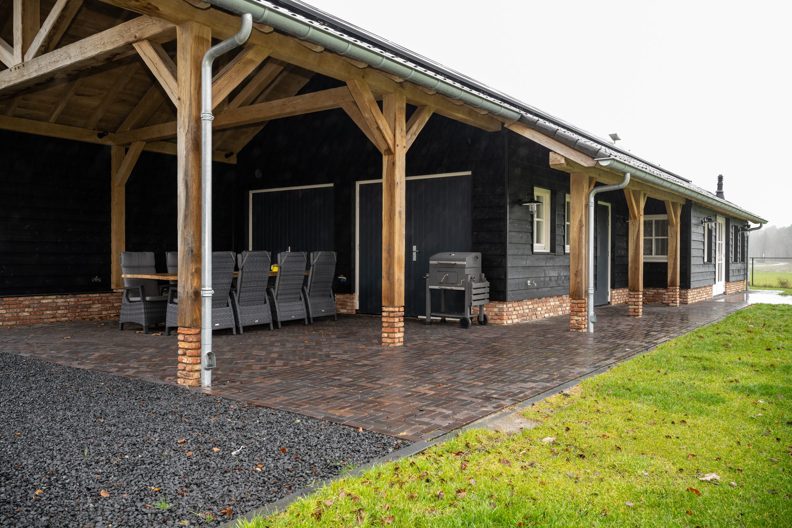 Vooraanzicht van een moderne barn-woning met zwarte houten gevelbekleding, witte kozijnen en een robuuste eikenhouten veranda op een stenen terras