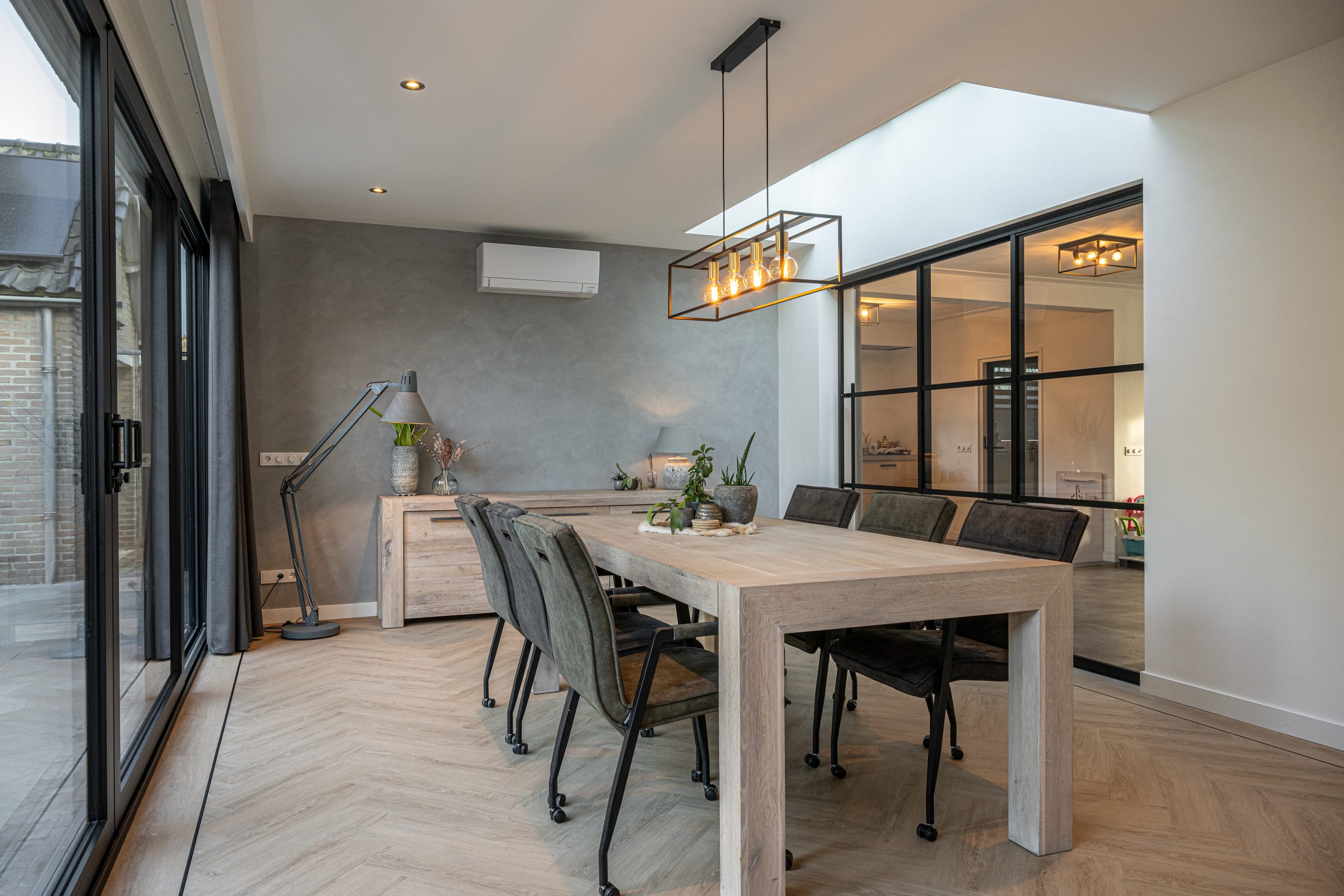 Moderne eetkamer met een grote houten tafel, een lichtstraat in het plafond en zwarte stalen deuren.