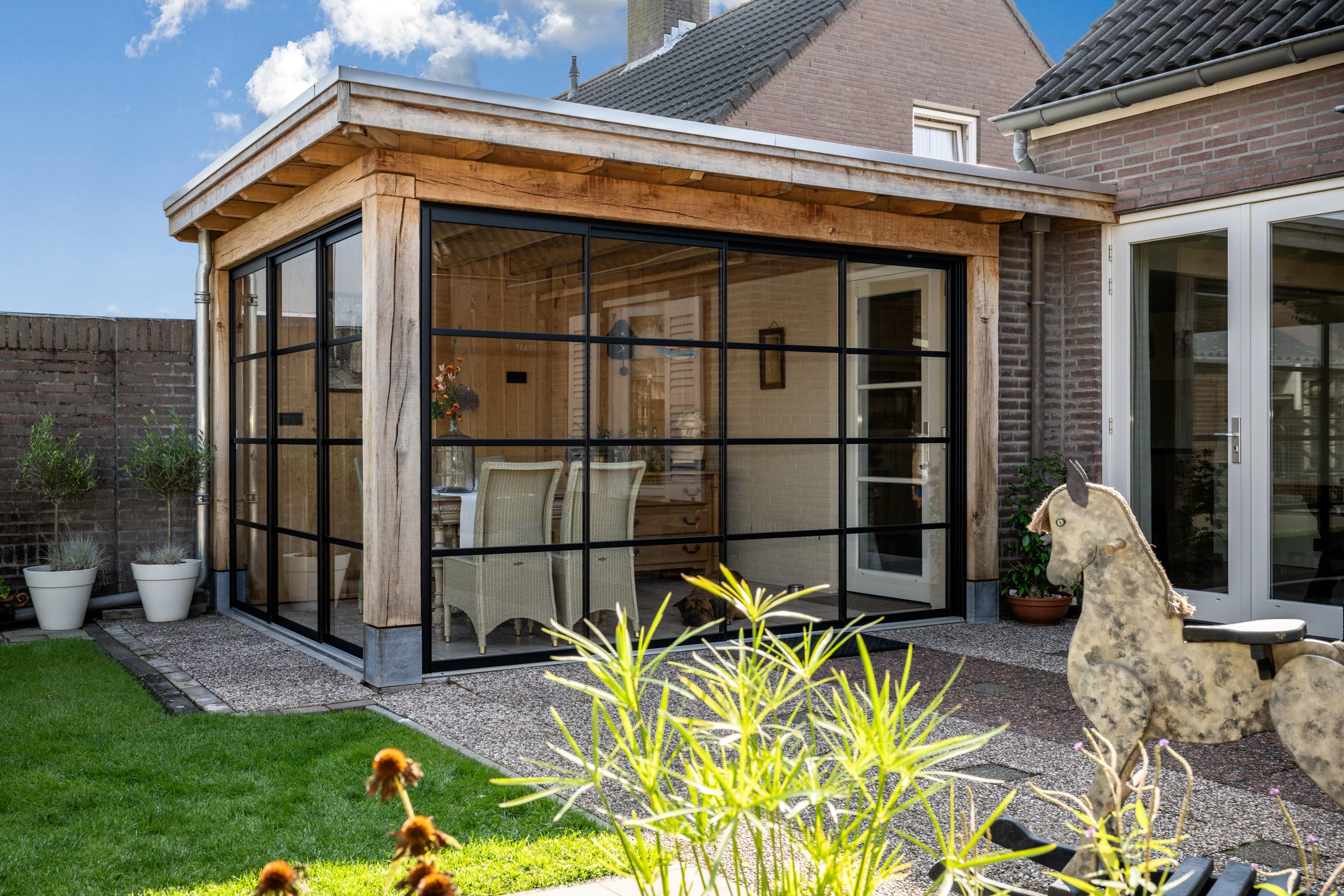 Een robuuste eikenhouten overkapping met zwarte stalen glaswanden in een achtertuin, ingericht met een eethoek en zicht op een stenen muur en beplanting.