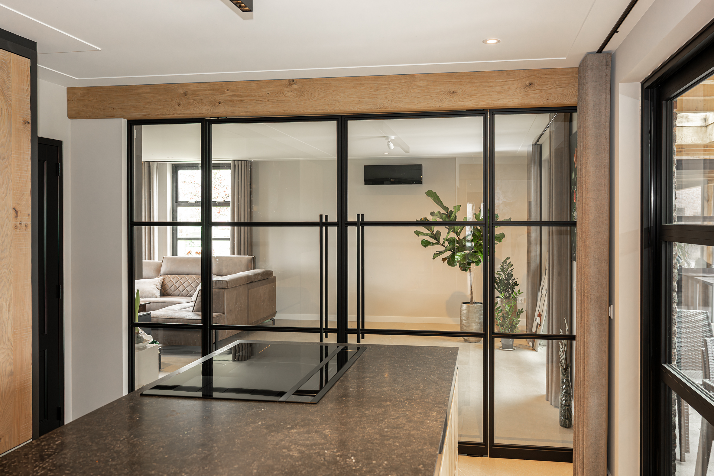 Zicht vanuit een moderne keuken door een zwarte stalen pui met glas naar een woonkamer met een grote kamerplant.