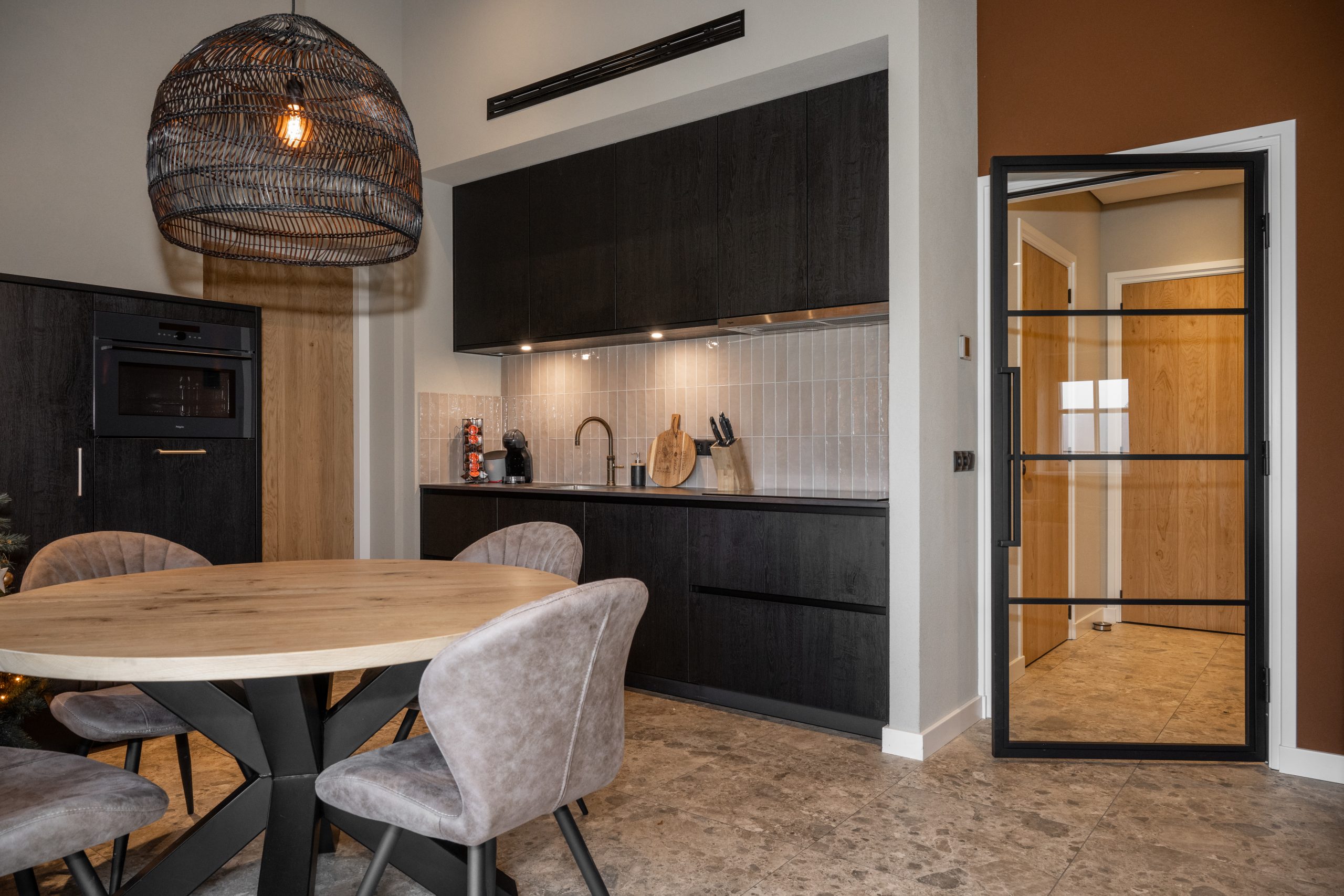 Modern interieur met een zwarte houten keuken, een ronde eiken eettafel en een zwarte stalen taatsdeur met glas.
