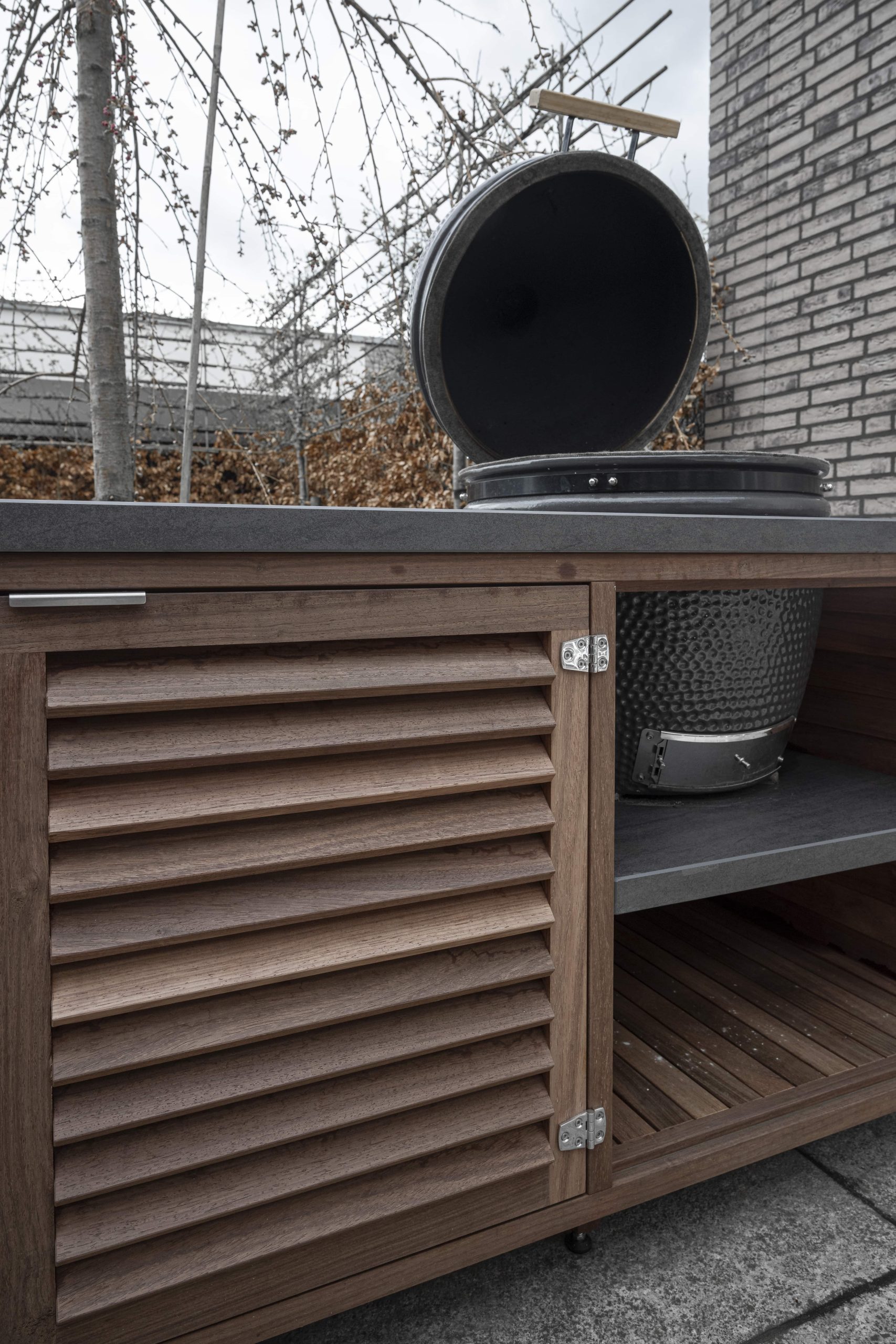 Detail van de louvre deurtjes en de binnenkant van een houten buitenkeuken met geopende barbecue.