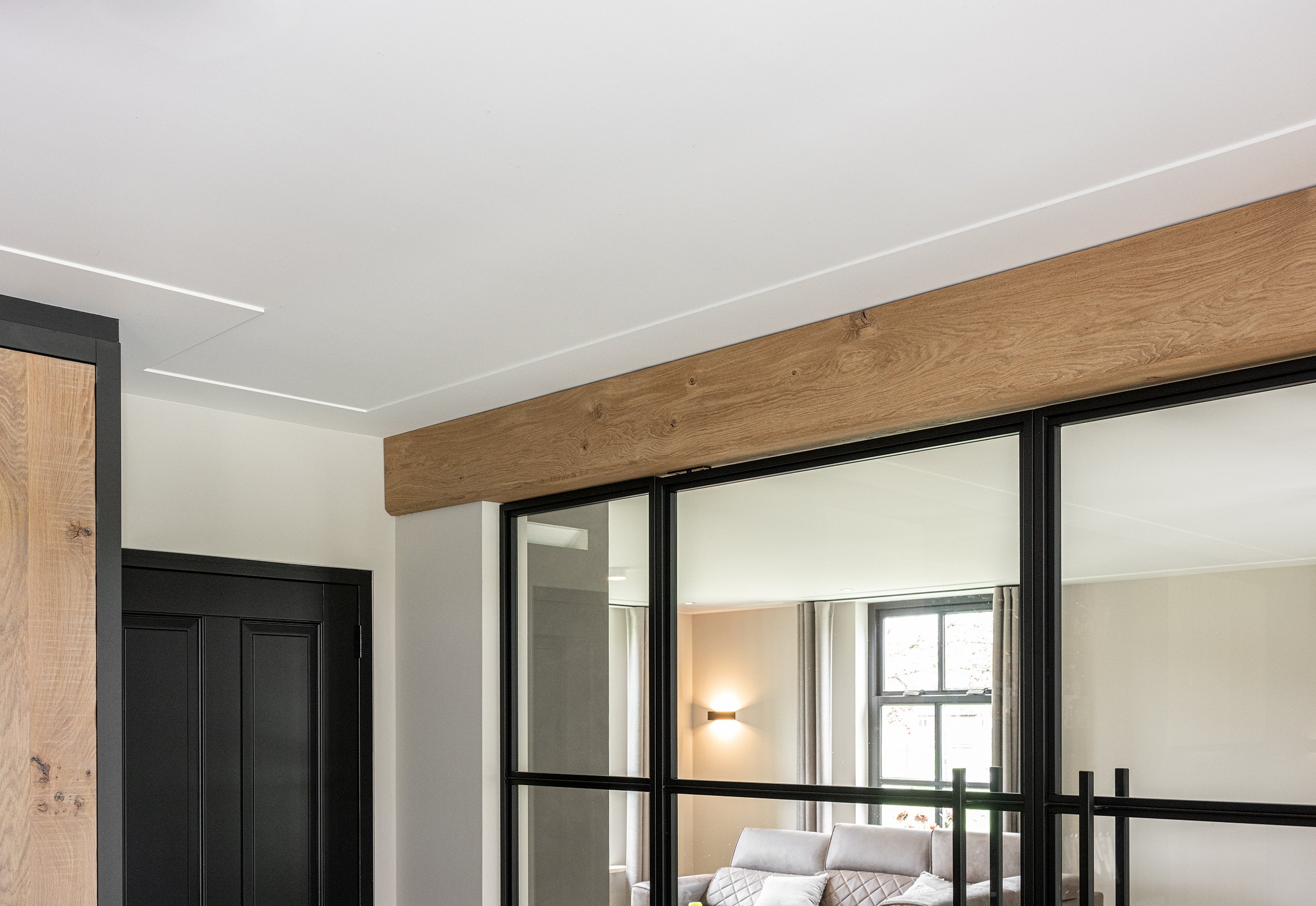 Bovenzijde van een zwarte stalen pui met glas, afgewerkt met een massieve eikenhouten balk tegen een wit plafond.