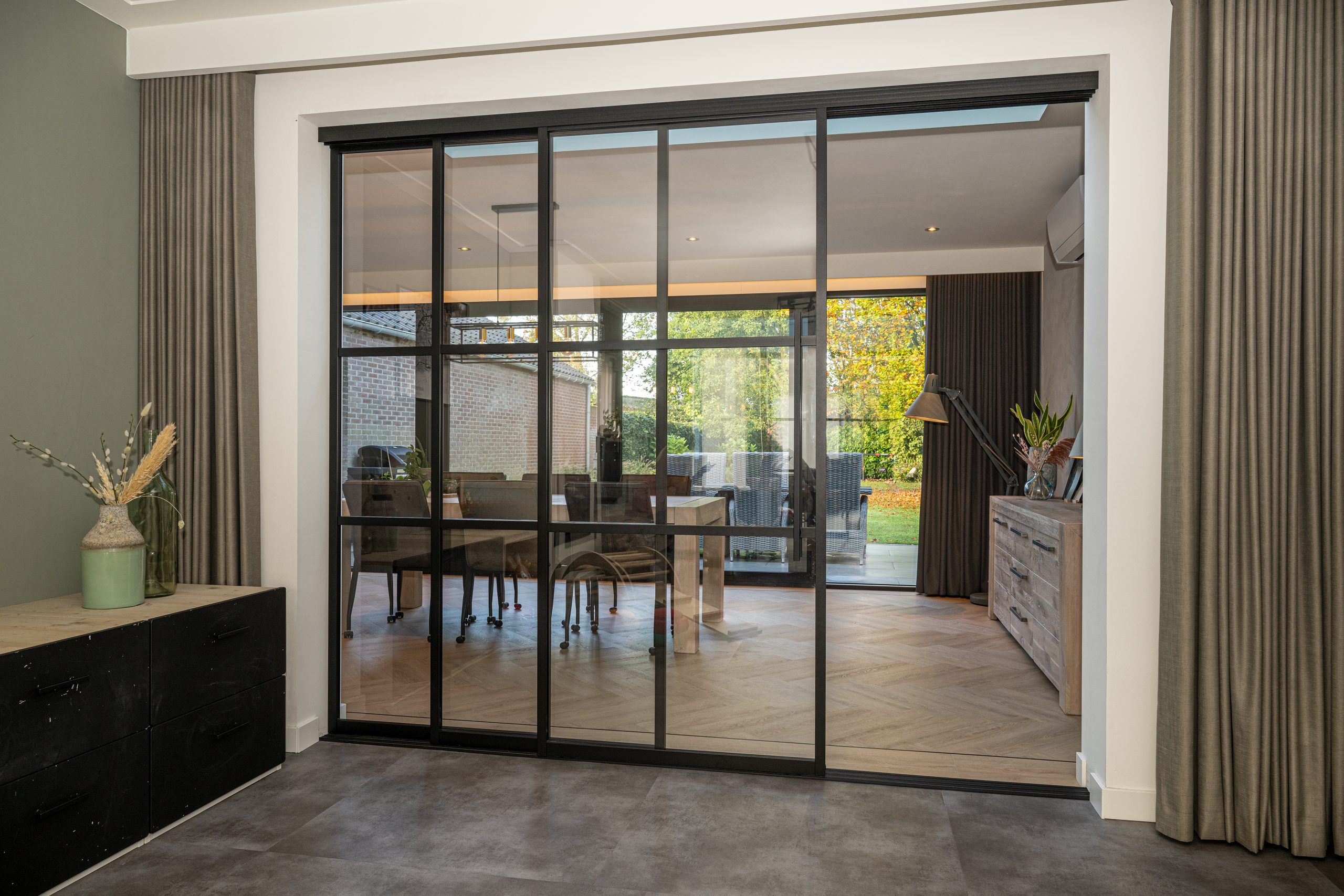 Zicht door gesloten zwarte stalen schuifdeuren naar een moderne eetkamer met visgraatvloer en tuinuitzicht.