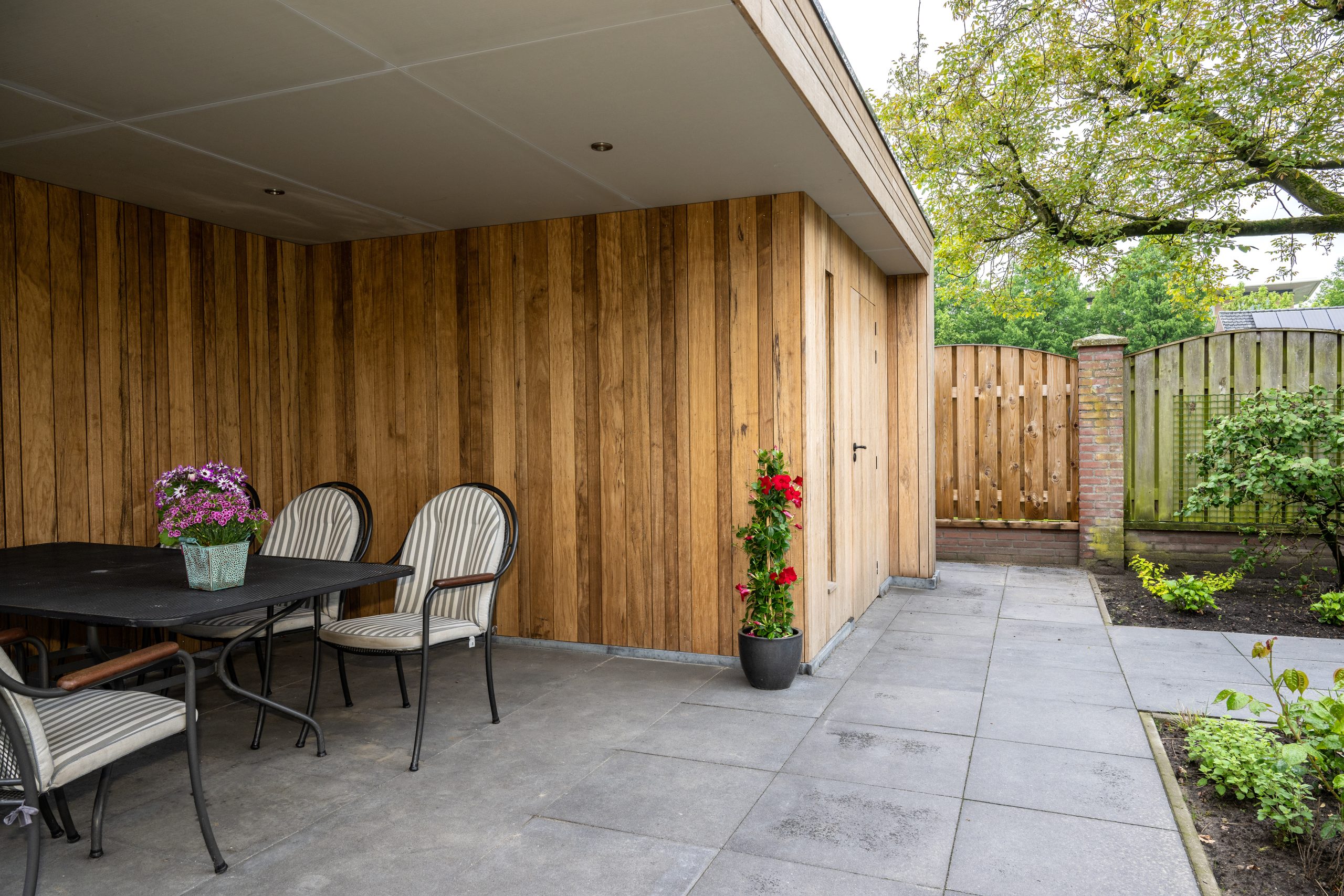 Interieur van een houten overkapping met inbouwspots in het plafond en zicht op een stenen schutting.