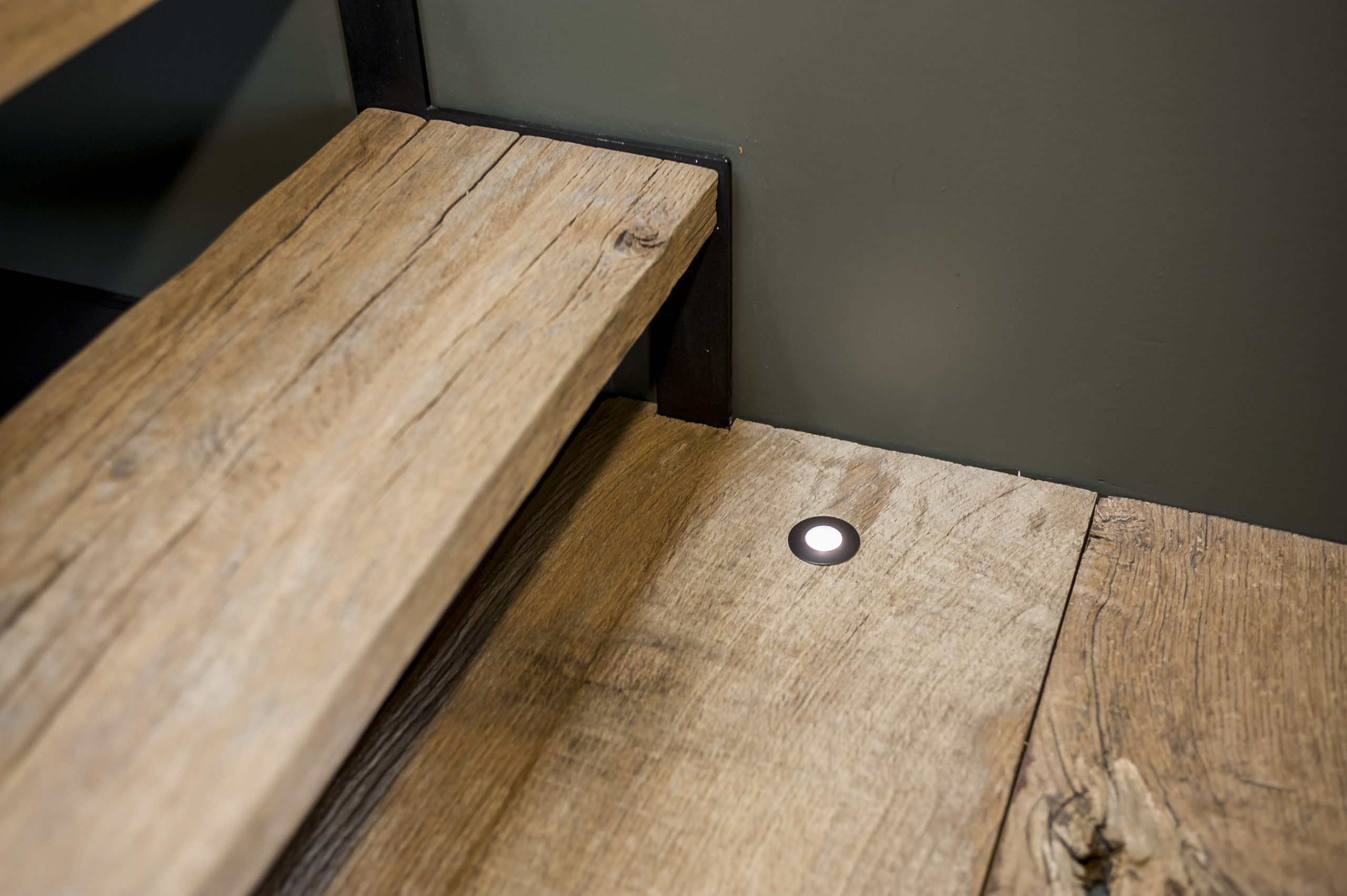 Detail van de aansluiting tussen een massief eiken trede, een zwart stalen frame en een verlichtingsspot.