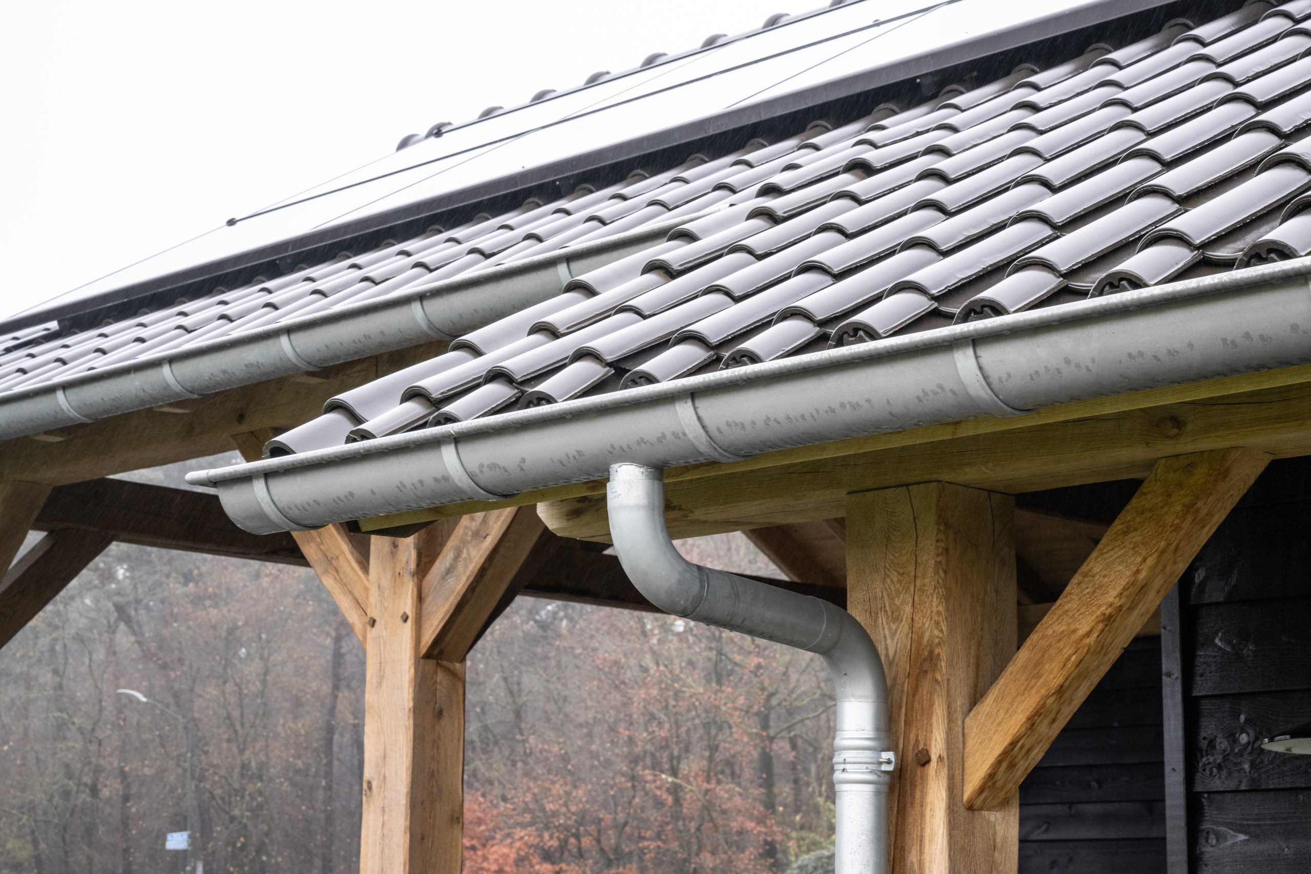 Detail van de dakafwerking met antraciet dakpannen, een zinken dakgoot en de zichtbare eikenhouten balken onder het overstek.
