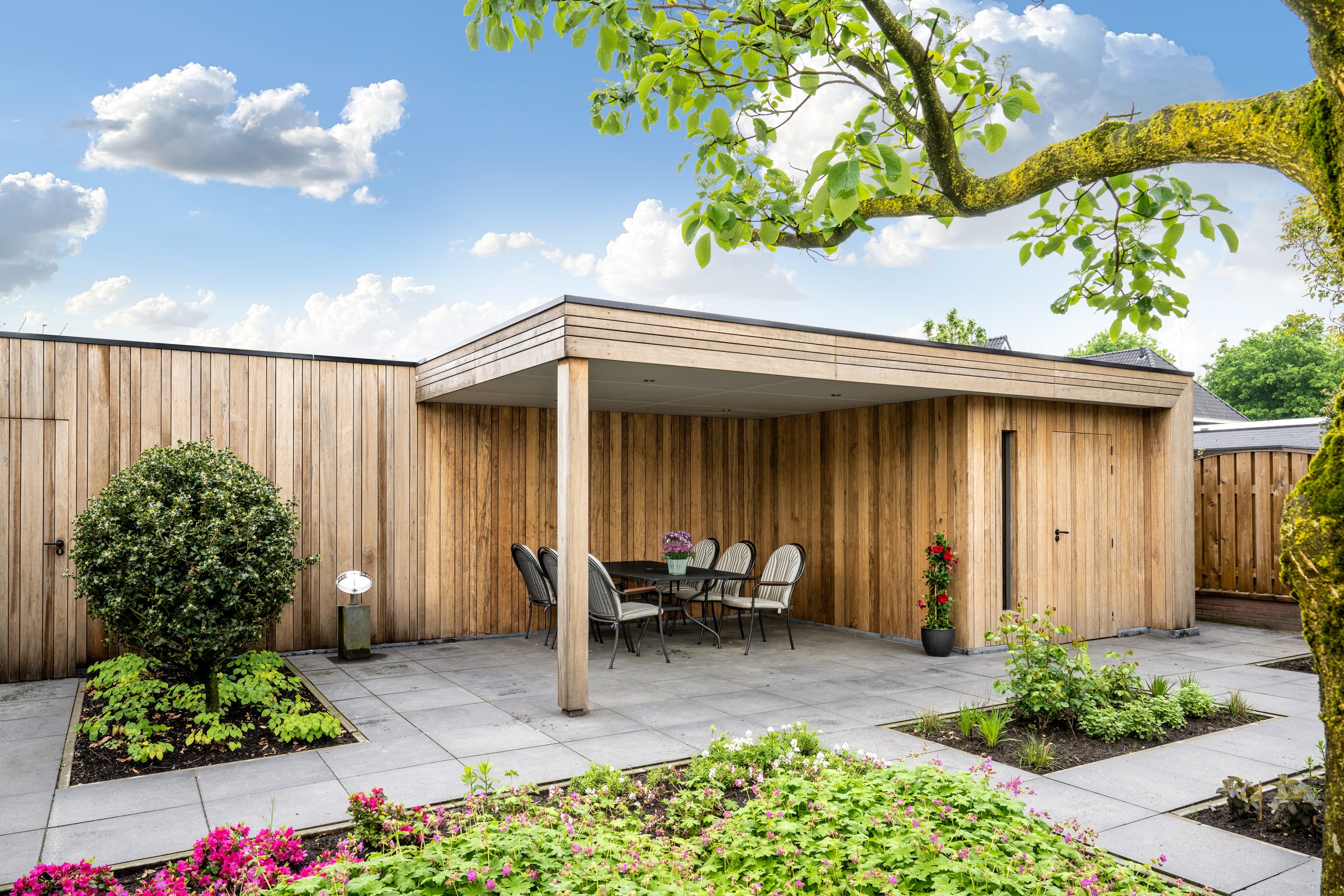 Een eethoek met gestreepte stoelen onder een houten overkapping, omgeven door borders met roze bloemen.