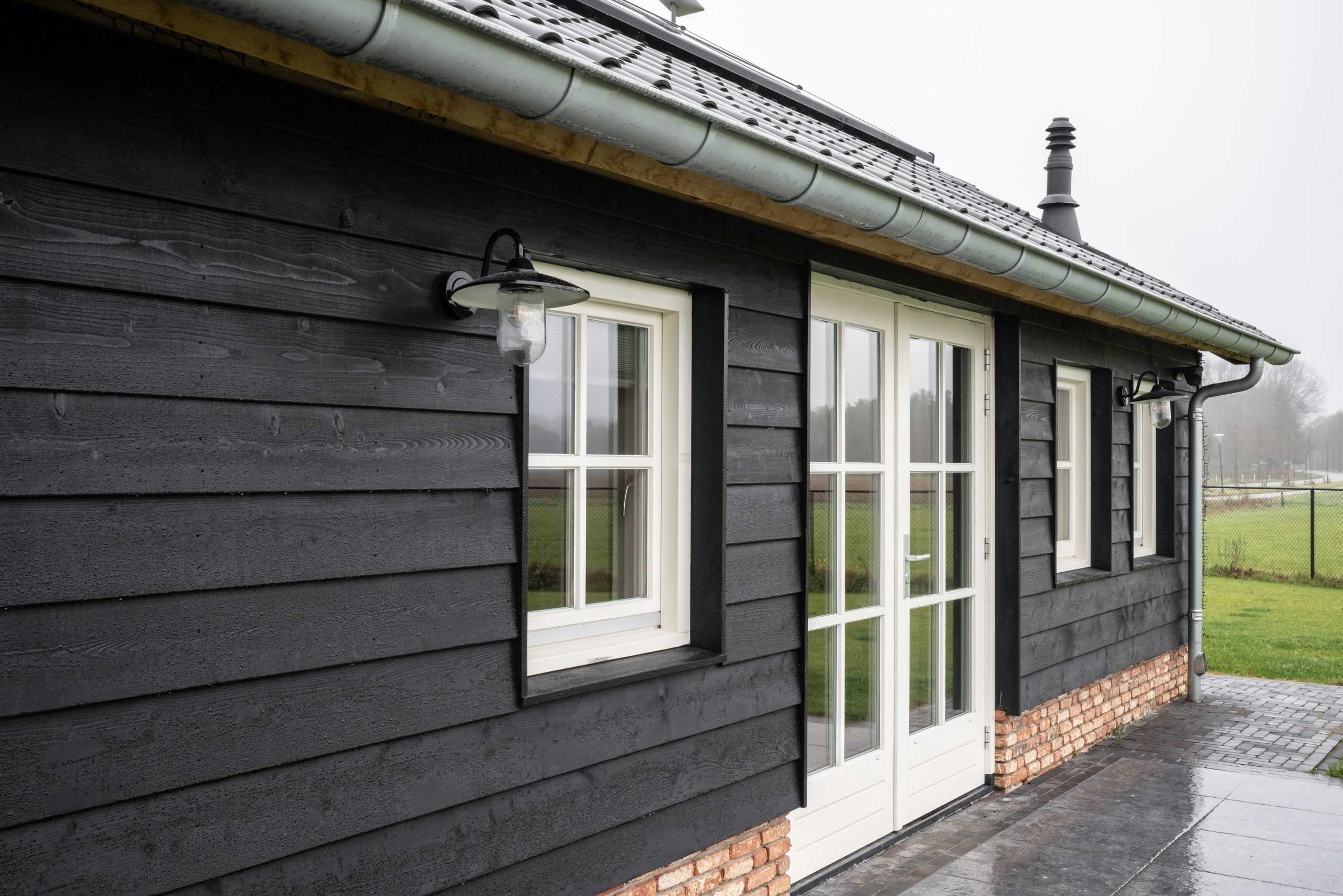 Close-up van een zwarte houten gevel met klassieke witte raamkozijnen en zwarte buitenverlichting van een luxe bijgebouw.