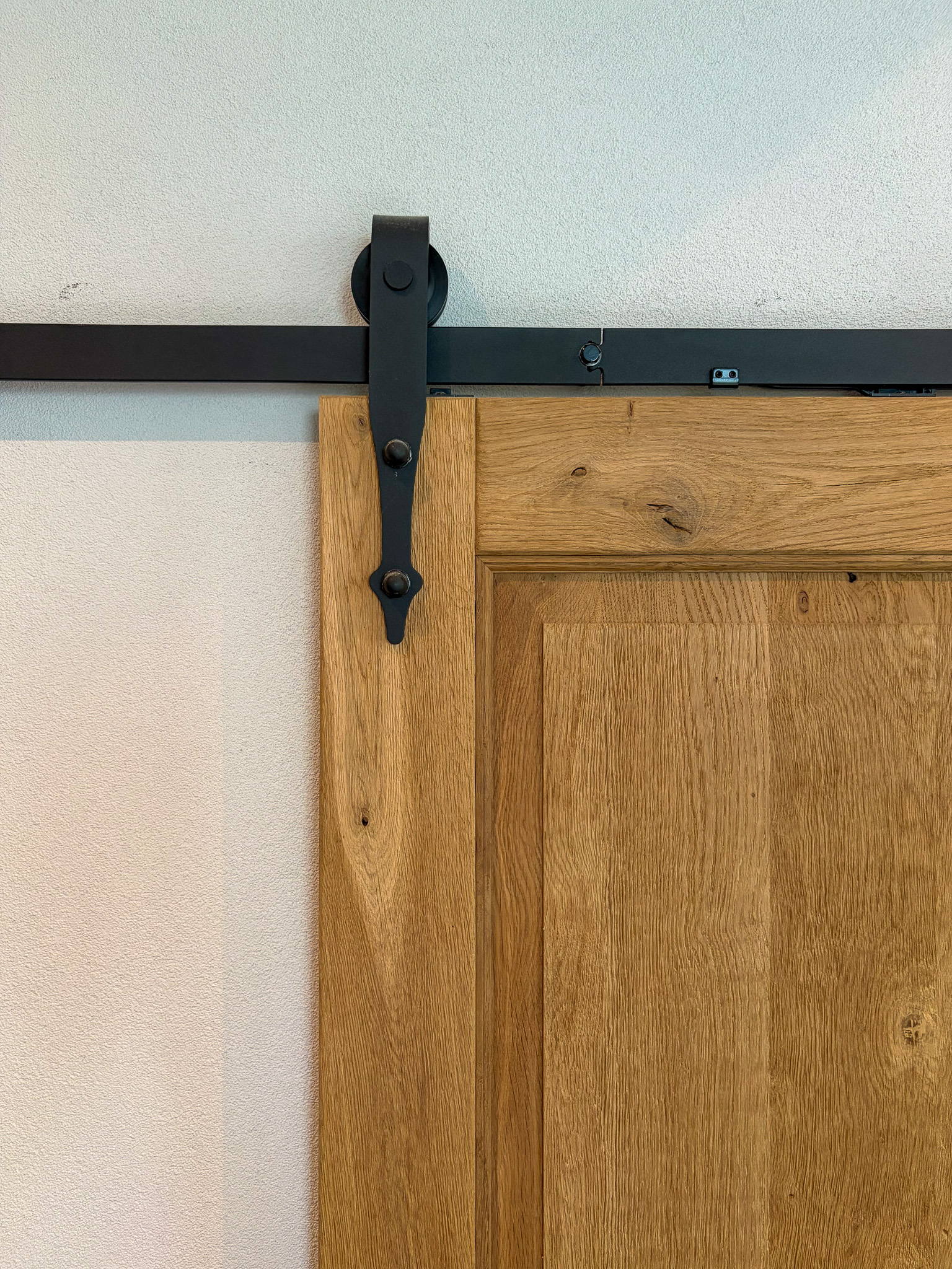 Close-up van een zwart stalen schuifdeurwiel en rail gemonteerd op een massief eikenhouten paneeldeur.