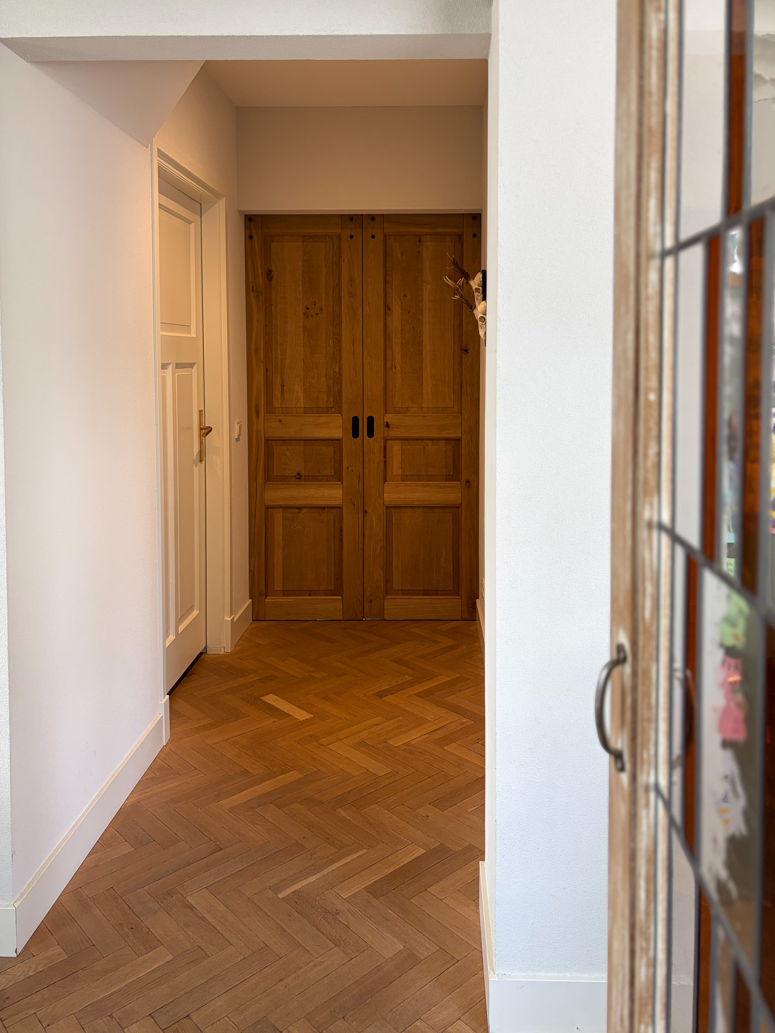 Doorkijk vanuit een kamer met glas-in-lood naar een gang met een visgraatvloer en eikenhouten schuifdeuren.