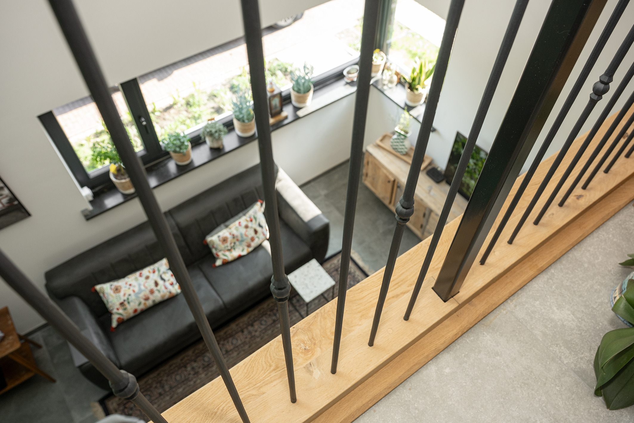 Uitzicht vanaf een vide naar beneden op een moderne woonkamer met een grijze bank en grote ramen, gezien door de spijlen van een trap.