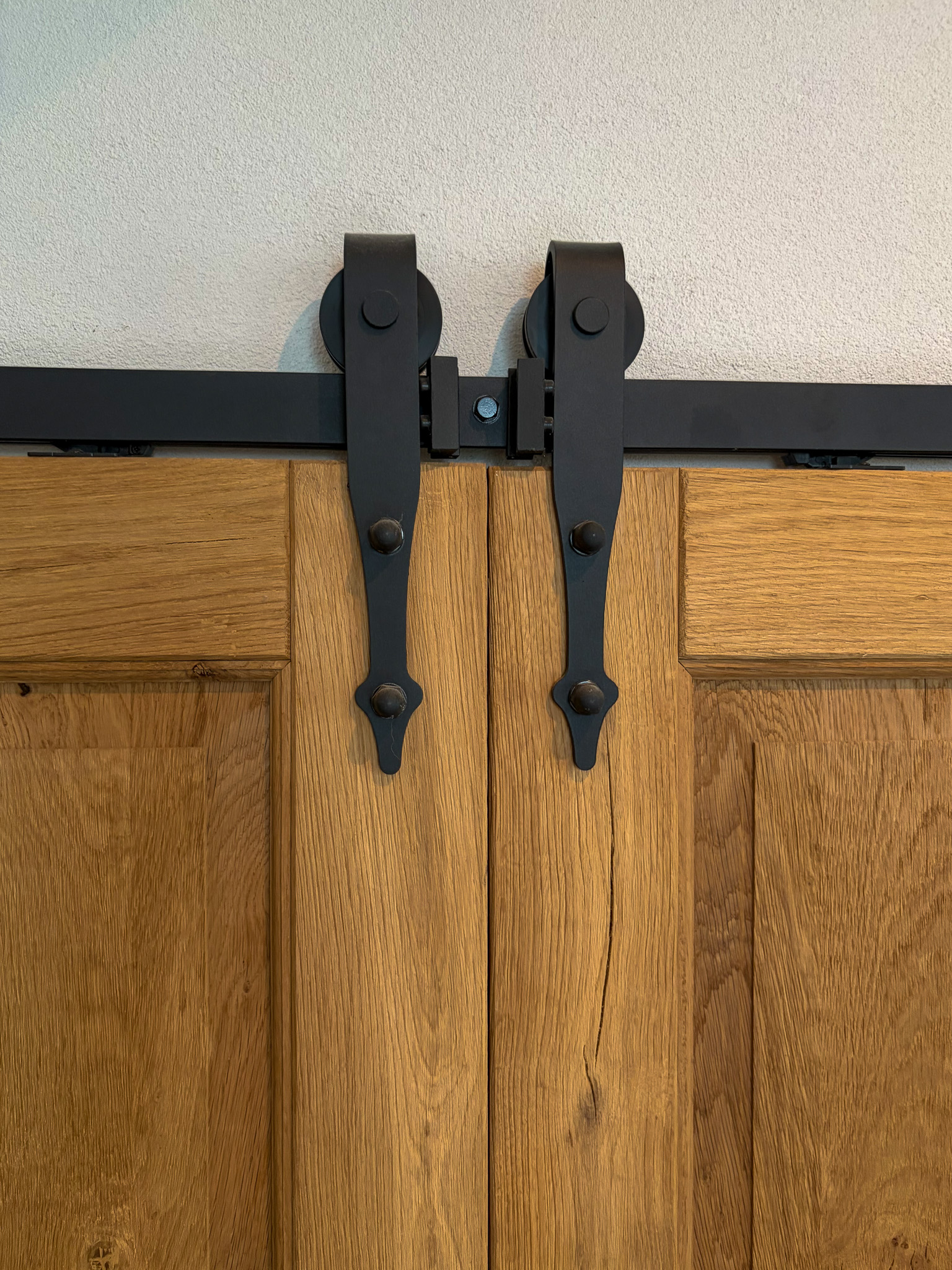 Een vooraanzicht van de bovenkant van twee massief eiken houten schuifdeuren met zwarte metalen rollersystemen en sierlijke beugels aan een zwarte rail.