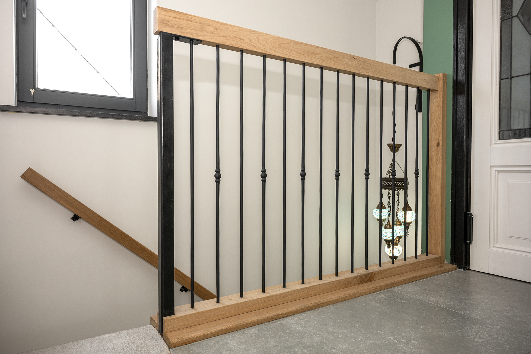 Moderne balustrade van eikenhout met slanke zwarte stalen spijlen en een hangende Turkse mozaïeklamp op de achtergrond.