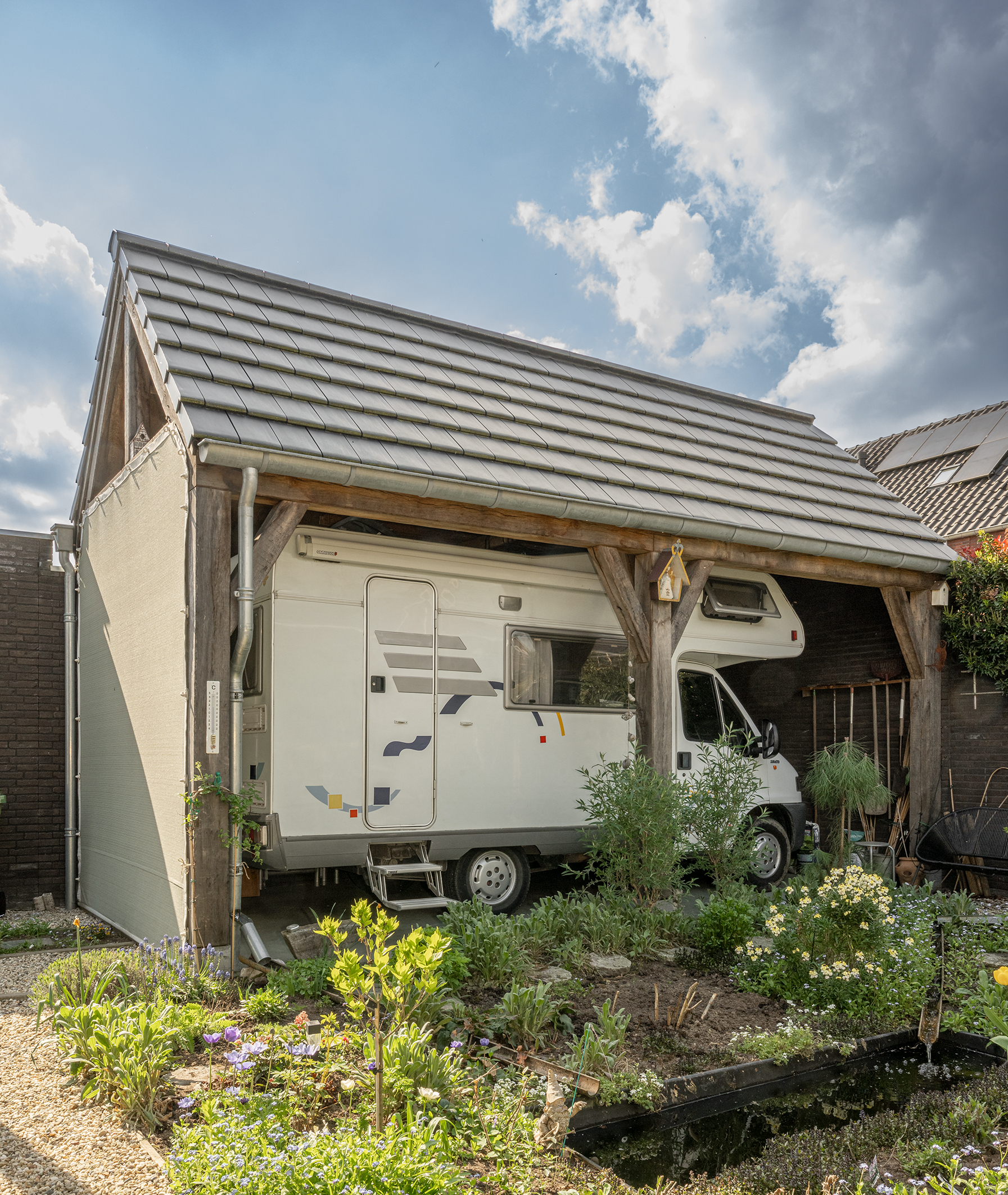 Een robuuste houten carport met een zadeldak van pannen, waaronder een witte camper geparkeerd staat in een tuin.