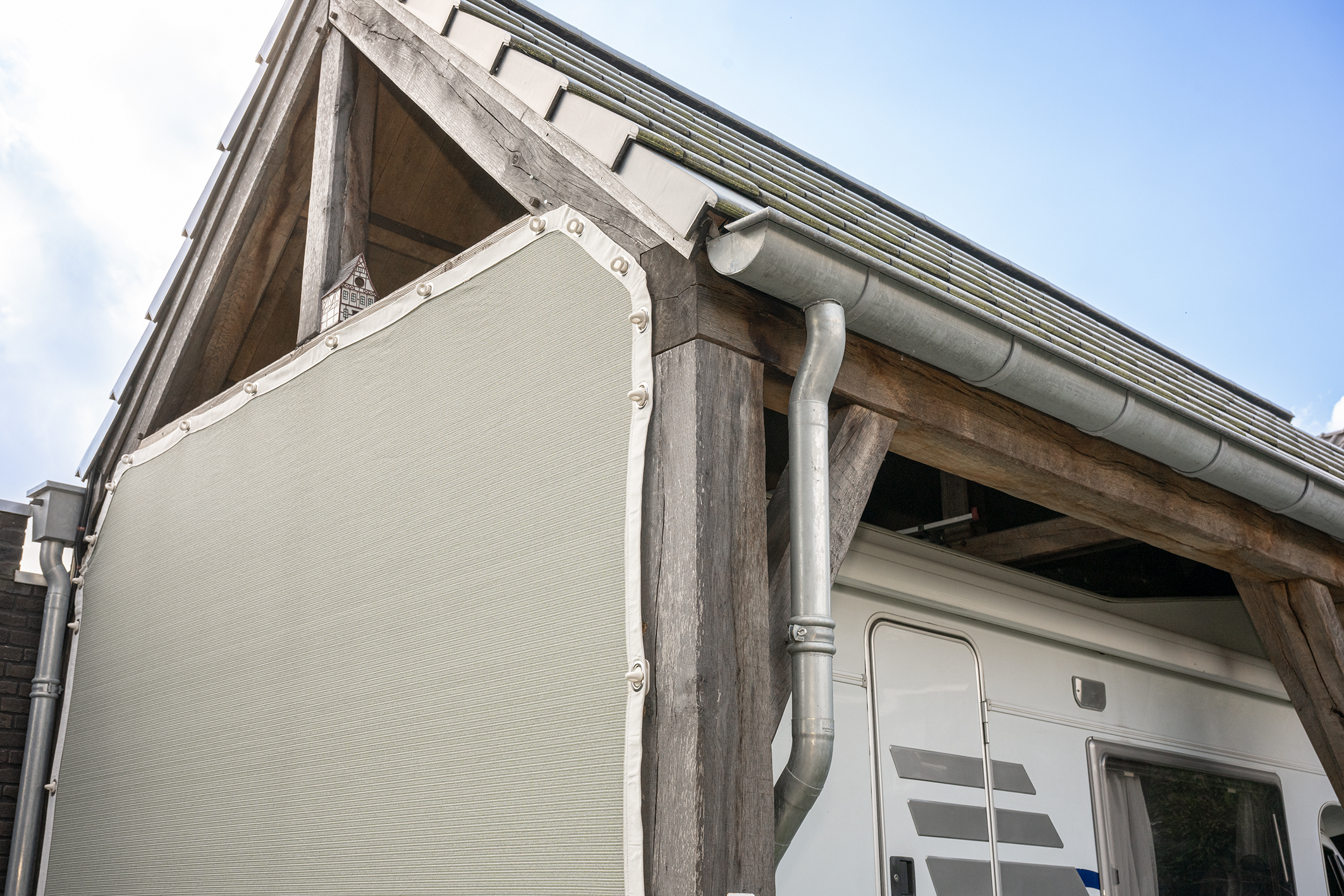 Een robuuste eikenhouten overkapping of carport met een zinken dakgoot, waaronder een witte camper geparkeerd staat.