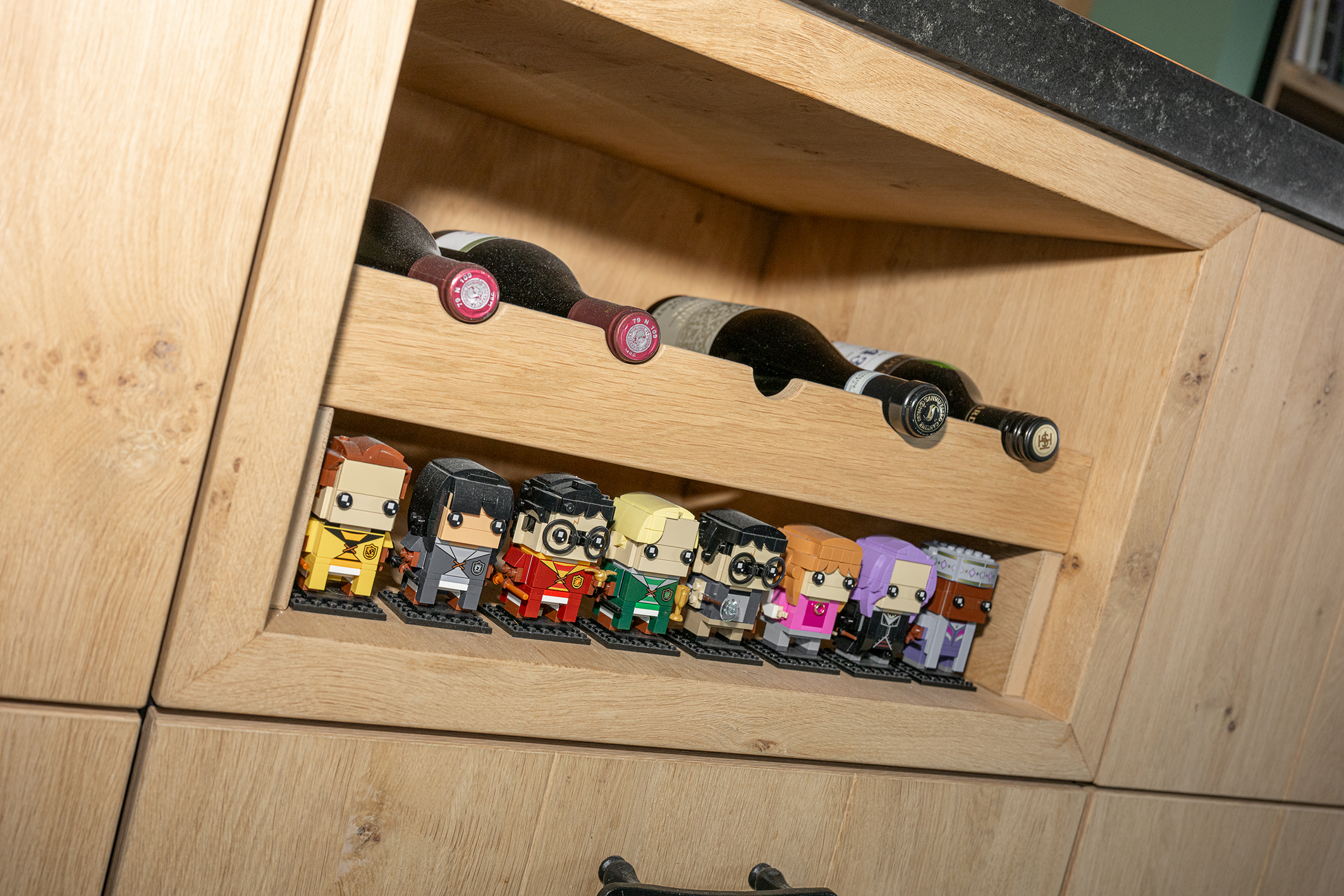 Een op maat gemaakt eikenhouten wijnrek in een keukeneiland, met daaronder een plank vol kleurrijke Harry Potter LEGO BrickHeadz figuren.