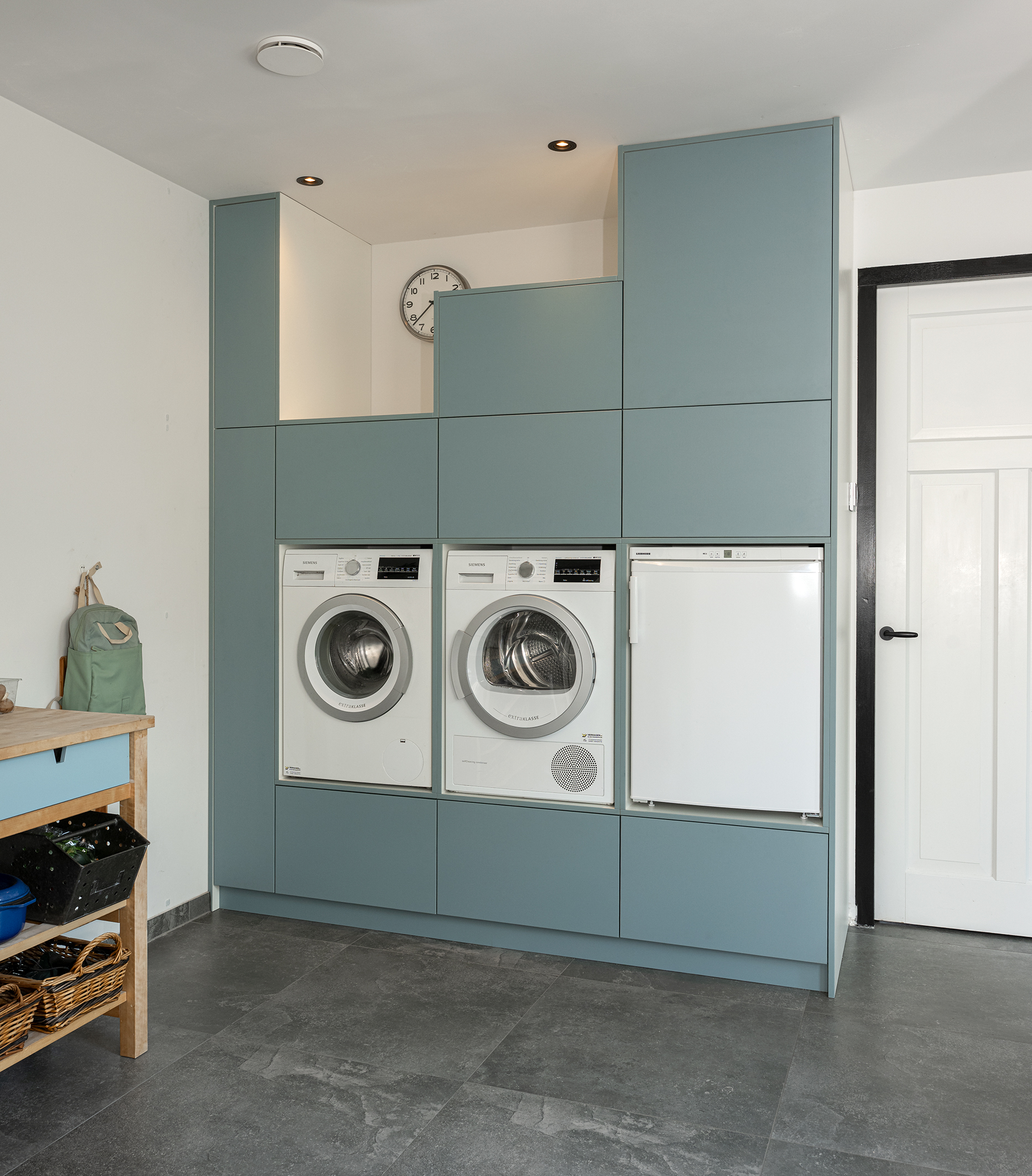 Ingebouwde maatwerkkast in een zachtblauwe kleur met nissen voor een wasmachine, droger en koelkast.