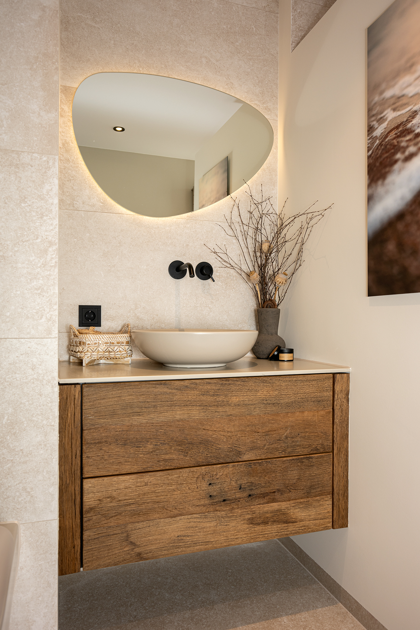 Modern houten wastafelmeubel met een beige opzetkom, een zwarte inbouwkraan en een organisch gevormde spiegel met achtergrondverlichting tegen een beige tegelwand.