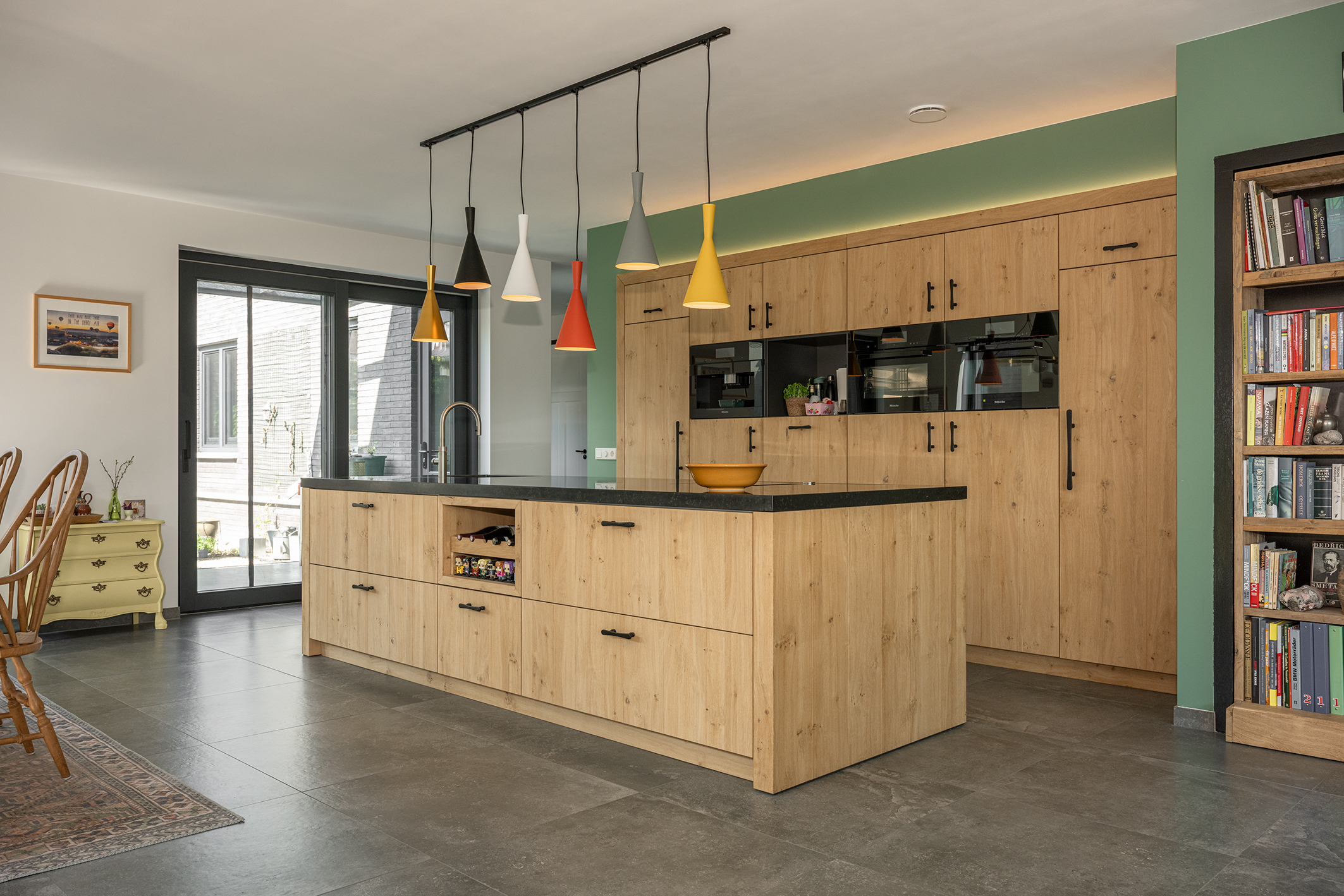 Ruime moderne keuken met eikenhouten kasten, een kookeiland met zwart blad en zes verschillend gekleurde hanglampen.