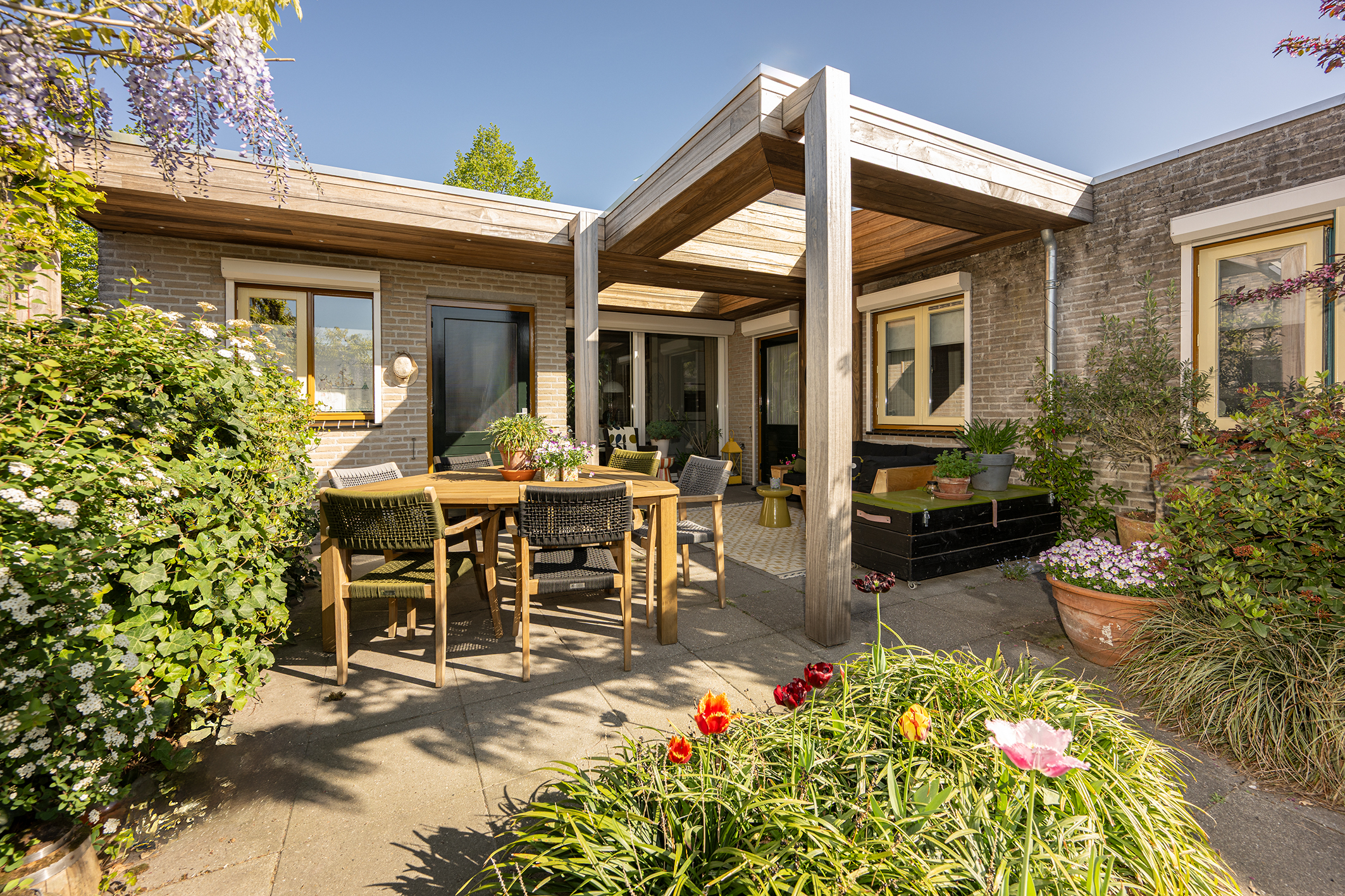 Overzicht van een zonnige tuin met een op maat gemaakte padoek houten overkapping die naadloos aansluit op de woning.