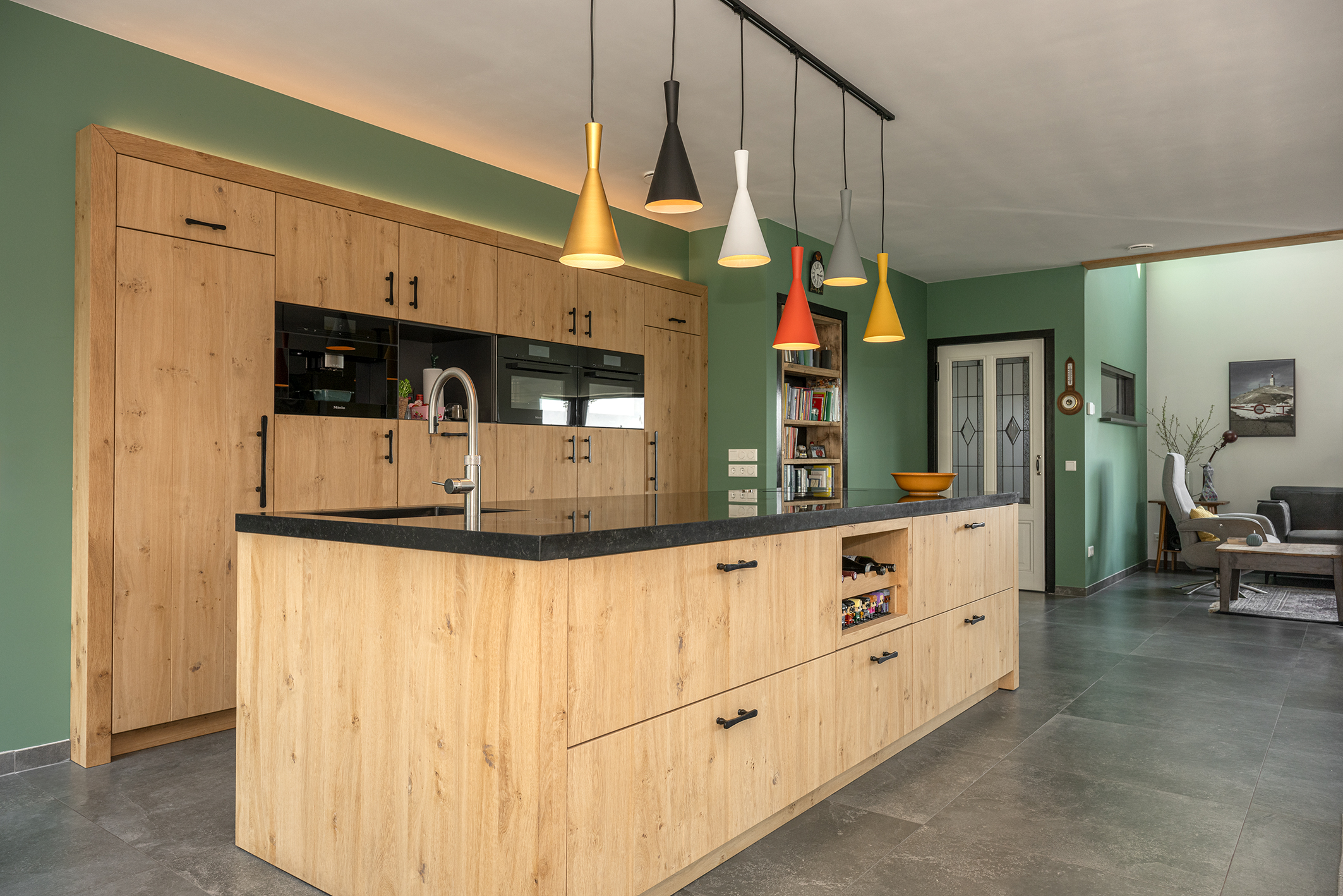 Overzicht van een open keuken die doorloopt in de woonkamer, met een antracietgrijze tegelvloer en veel natuurlijk licht.