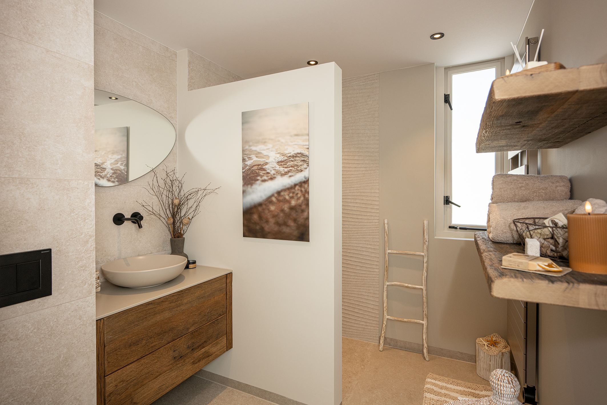 Overzicht van een lichte badkamer met een eikenhouten wastafelmeubel, een witte tussenmuur met kunstwerk en een doorkijk naar de inloopdouche met een houten ladder.
