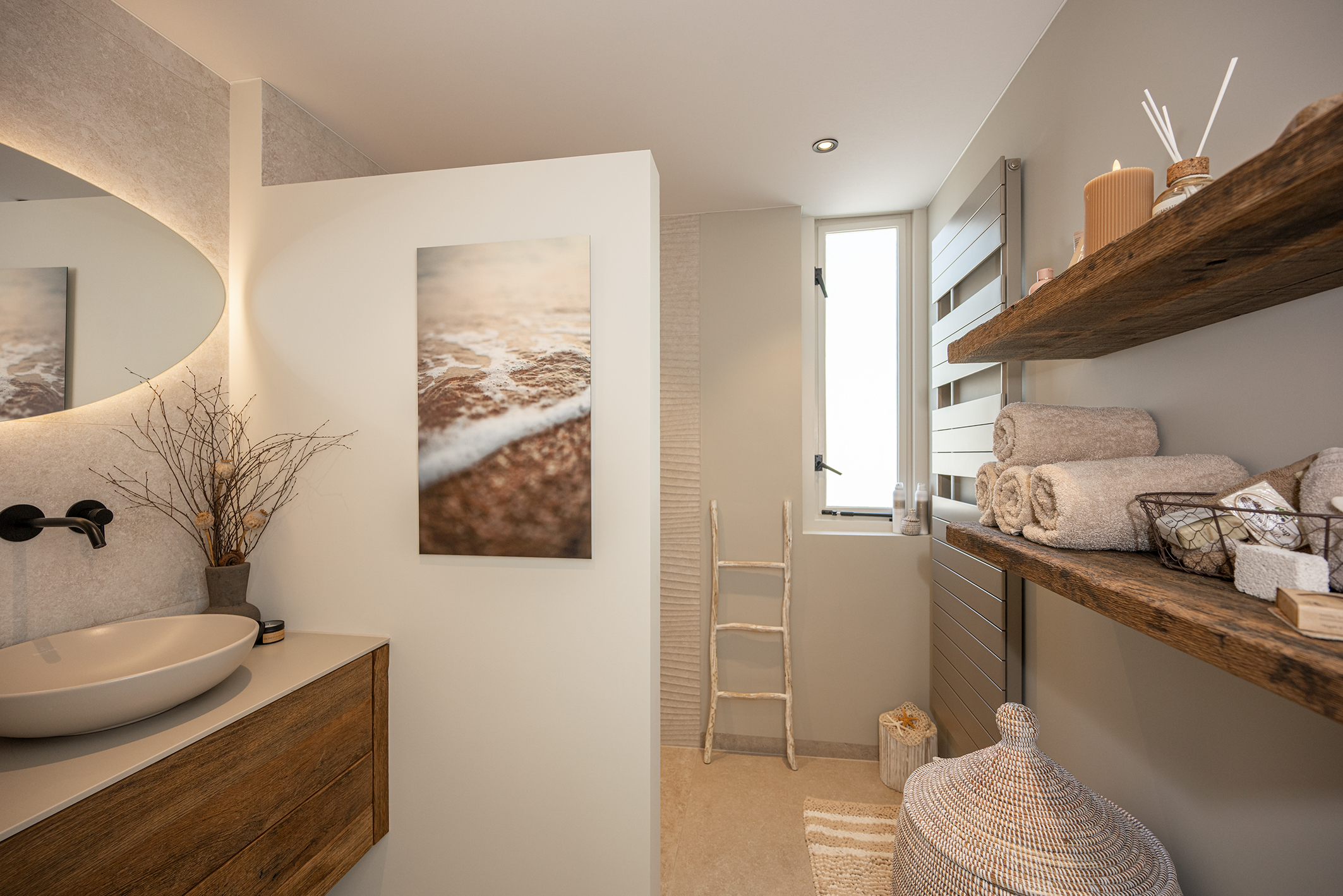 Een ruimtelijk beeld van de badkamer waarin het houten wastafelmeubel, de verlichte organische spiegel en de houten wandschappen met accessoires samenkomen voor een rustieke uitstraling.