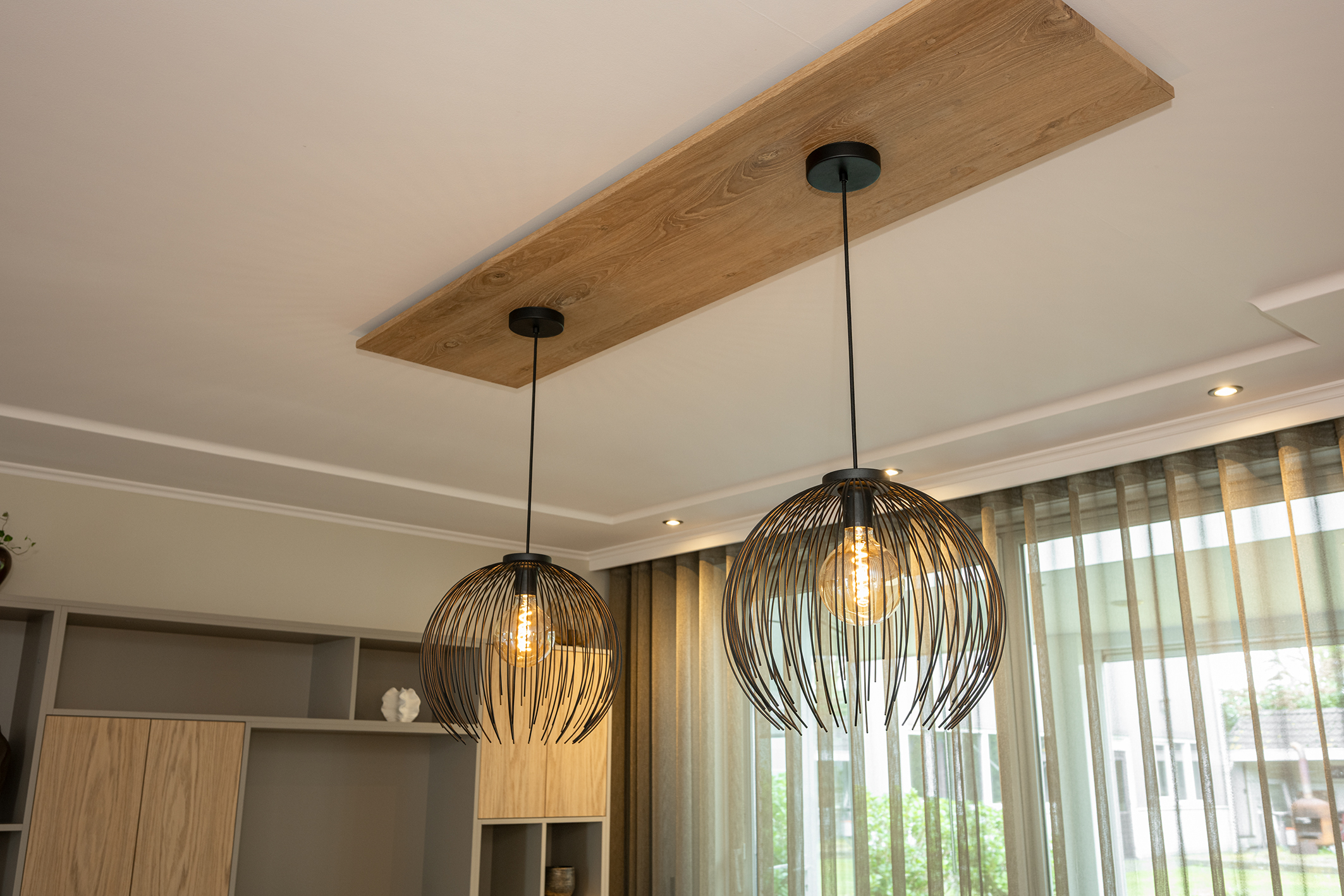 Twee zwarte draadstalen hanglampen gemonteerd aan een eikenhouten plafondpaneel boven een eettafel.