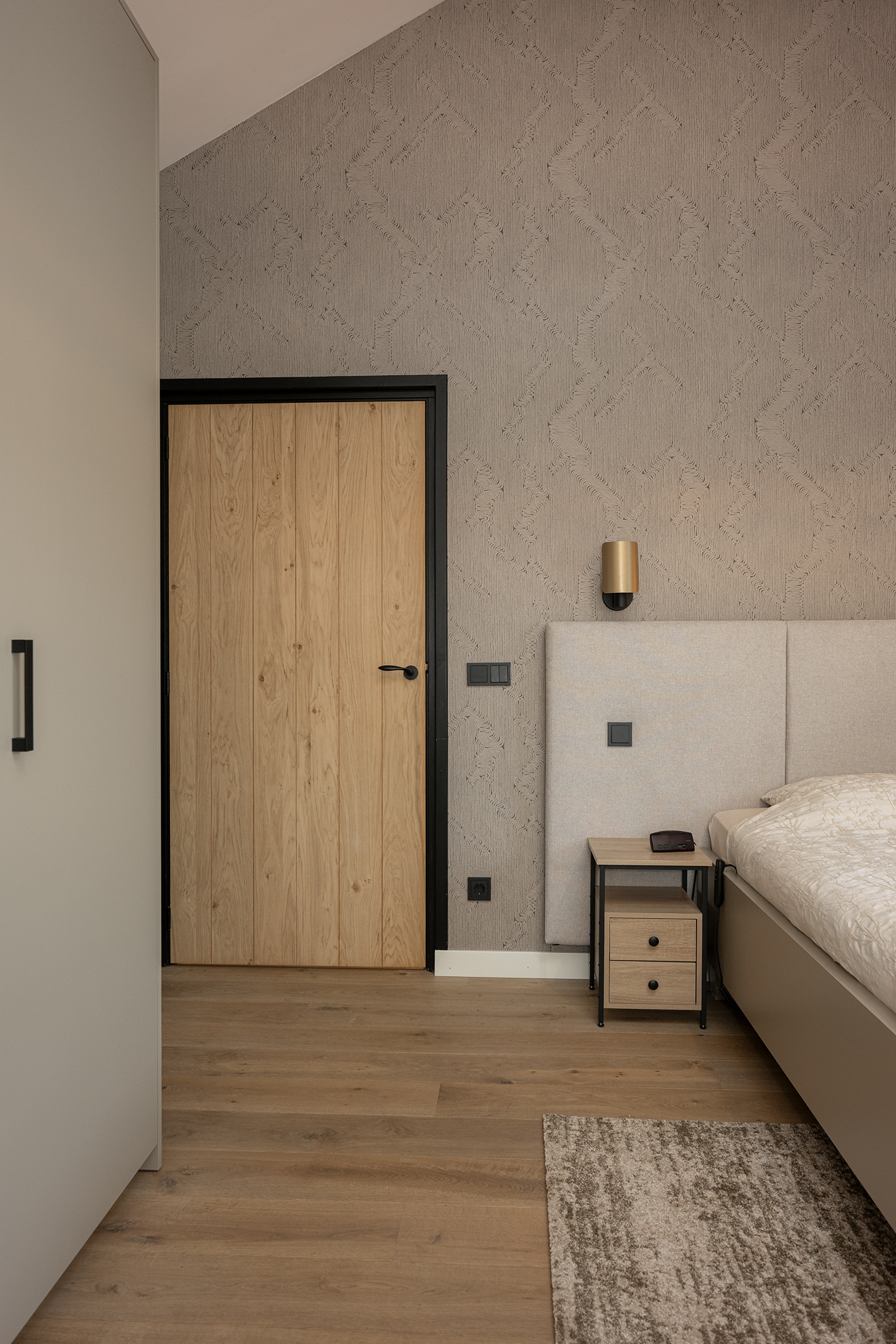 Een gouden wandlamp boven een nachtkastje in de slaapkamer, gemonteerd op een muur met een golvend 3D-patroon behang.