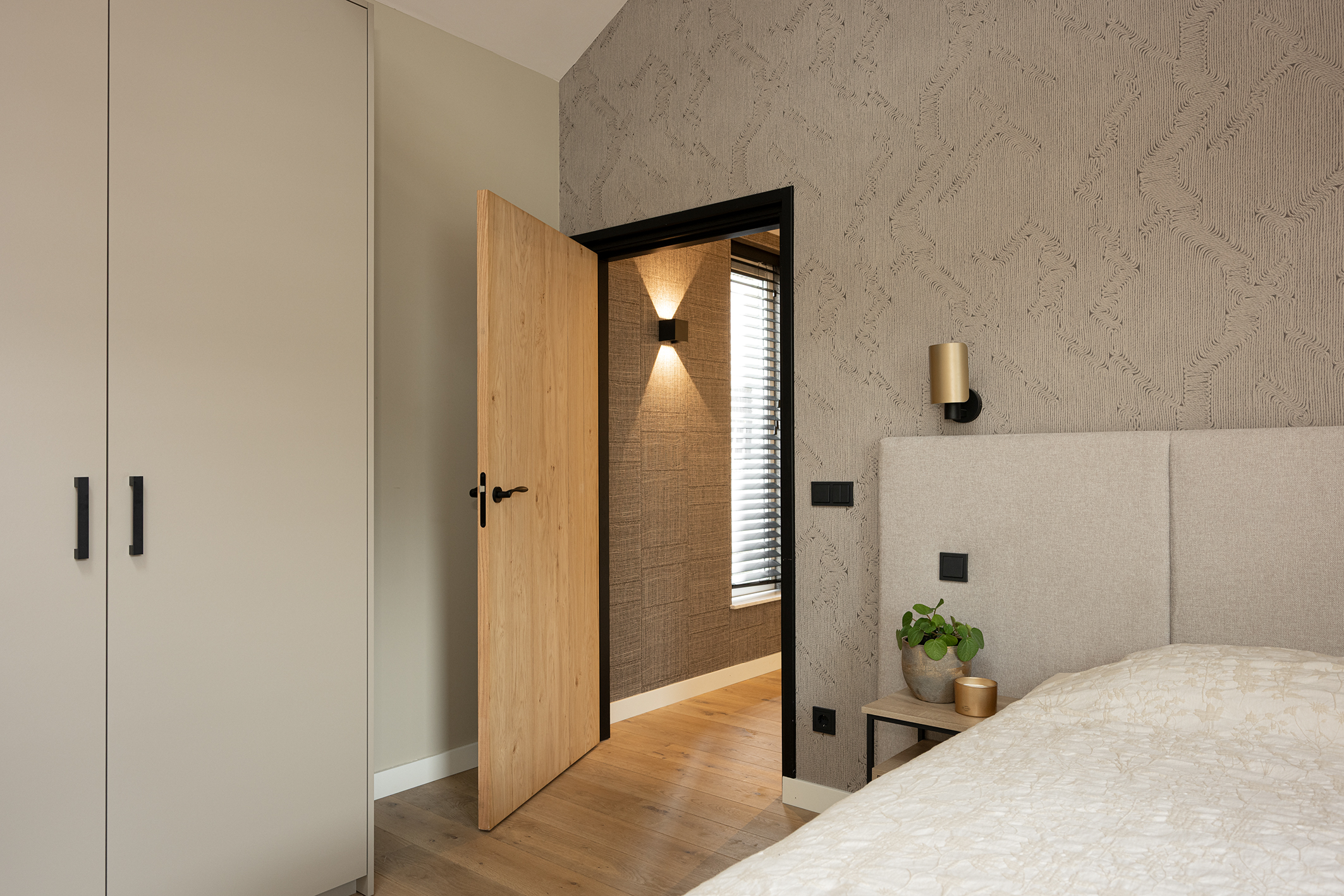 Zicht vanuit de slaapkamer door een openstaande houten deur naar een hal met donker geweven behang en moderne wandverlichting.