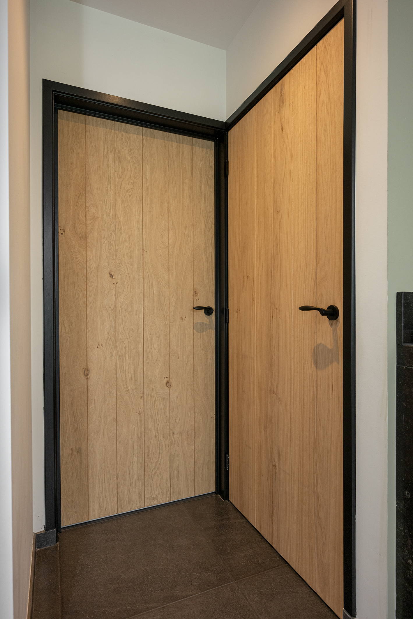 Twee moderne eikenhouten binnendeuren met verticale nerven en zwarte deurklinken in een zwart kozijn.