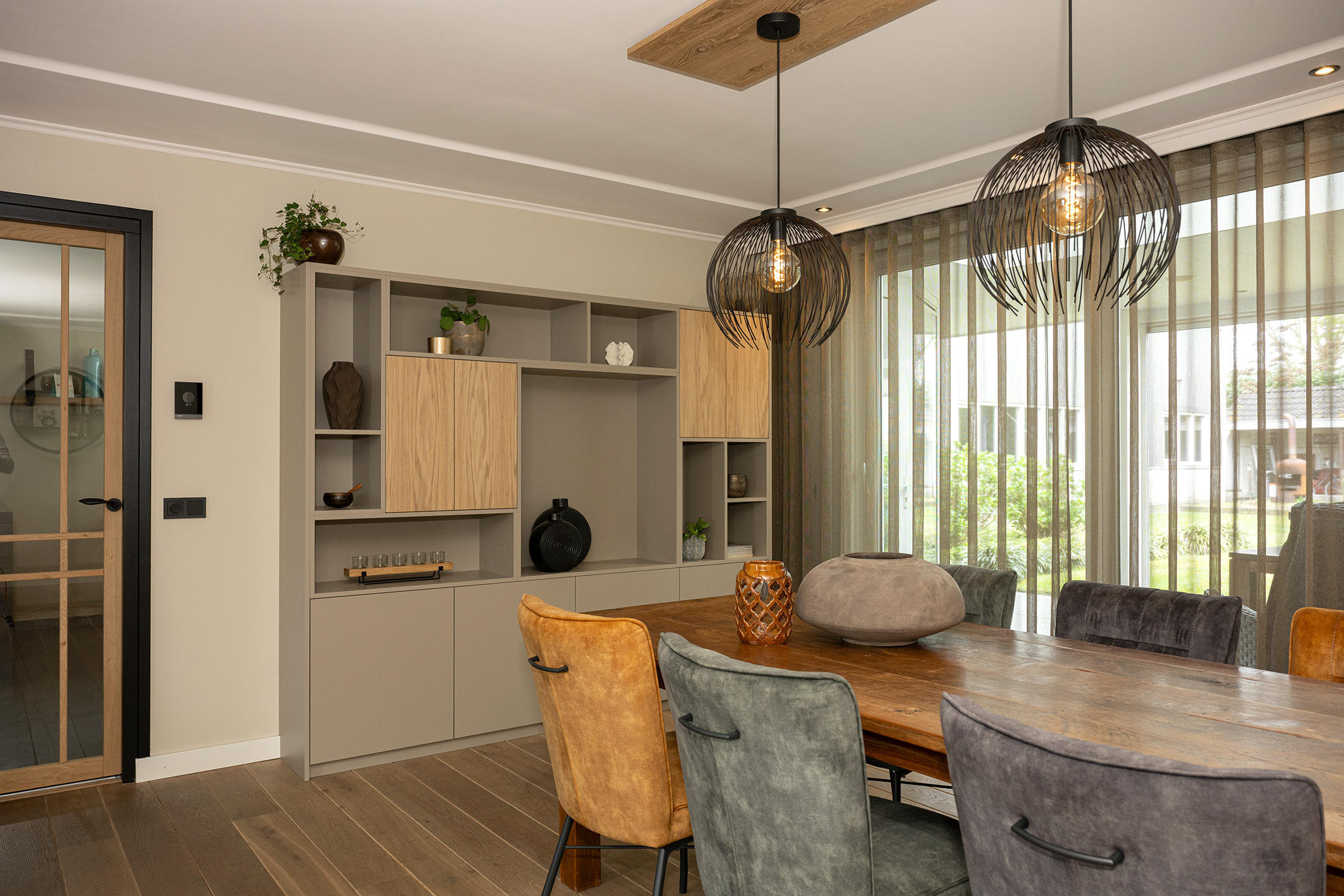 Ruime eethoek met een lange robuuste houten tafel, moderne eetkamerstoelen, twee grote zwarte hanglampen en een grijze maatwerkkast.