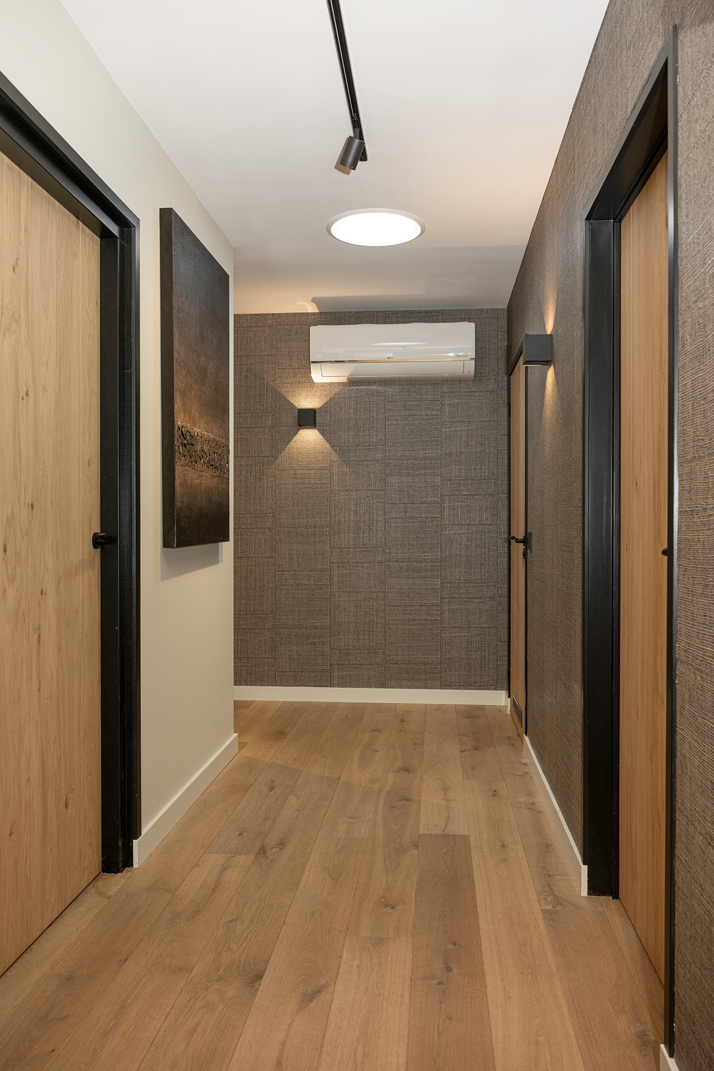 Moderne gang met een warme houten vloer, donker textielbehang van Arte, strakke wandverlichting en houten binnendeuren.