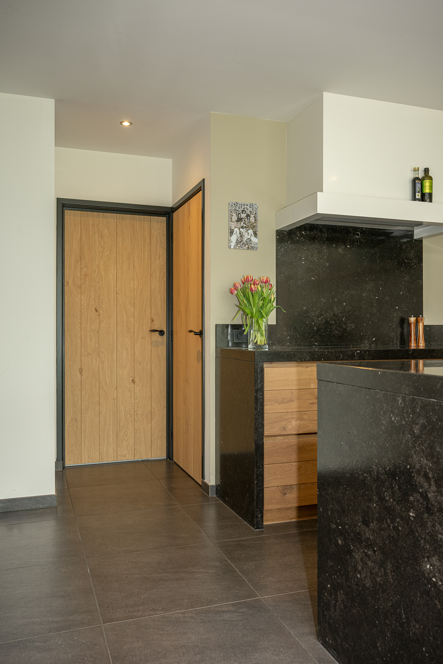 Een gedetailleerde, lage perspectiefopname vanuit de keuken, gericht op de hal. In beeld zijn kamerhoge houten binnendeuren met strakke zwarte kozijnen en handgrepen. Op de voorgrond zijn de gootsteen en een vaas rode tulpen zichtbaar.