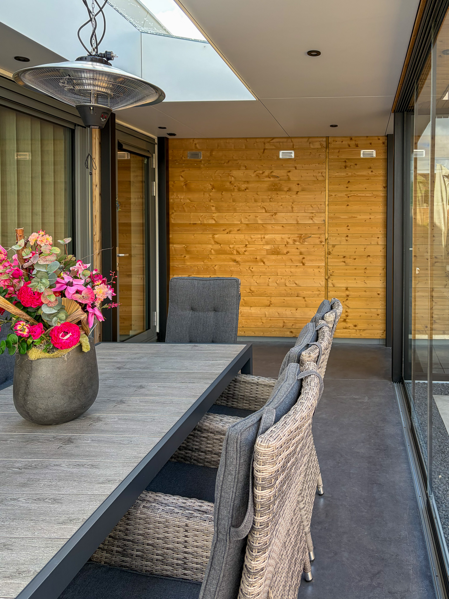Het interieur van de overkapping met een houten eettafel, een kleurrijk boeket bloemen en een terrasverwarmer aan het plafond.