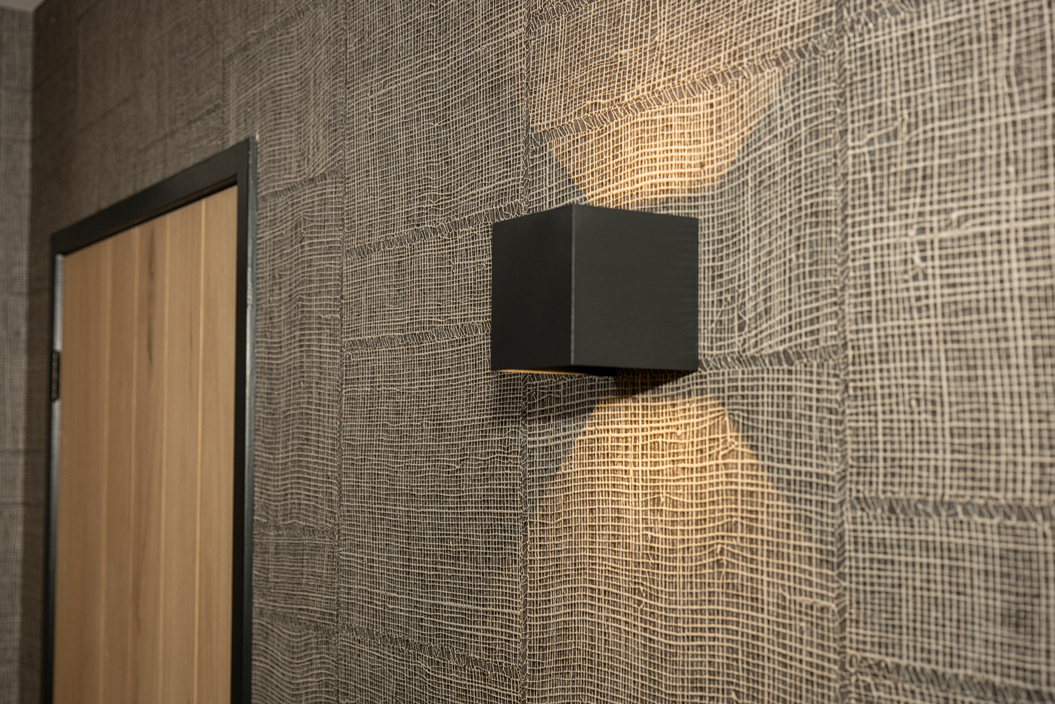 Een moderne zwarte kubusvormige wandlamp die sfeervol licht werpt op grof geweven structuurbehang.