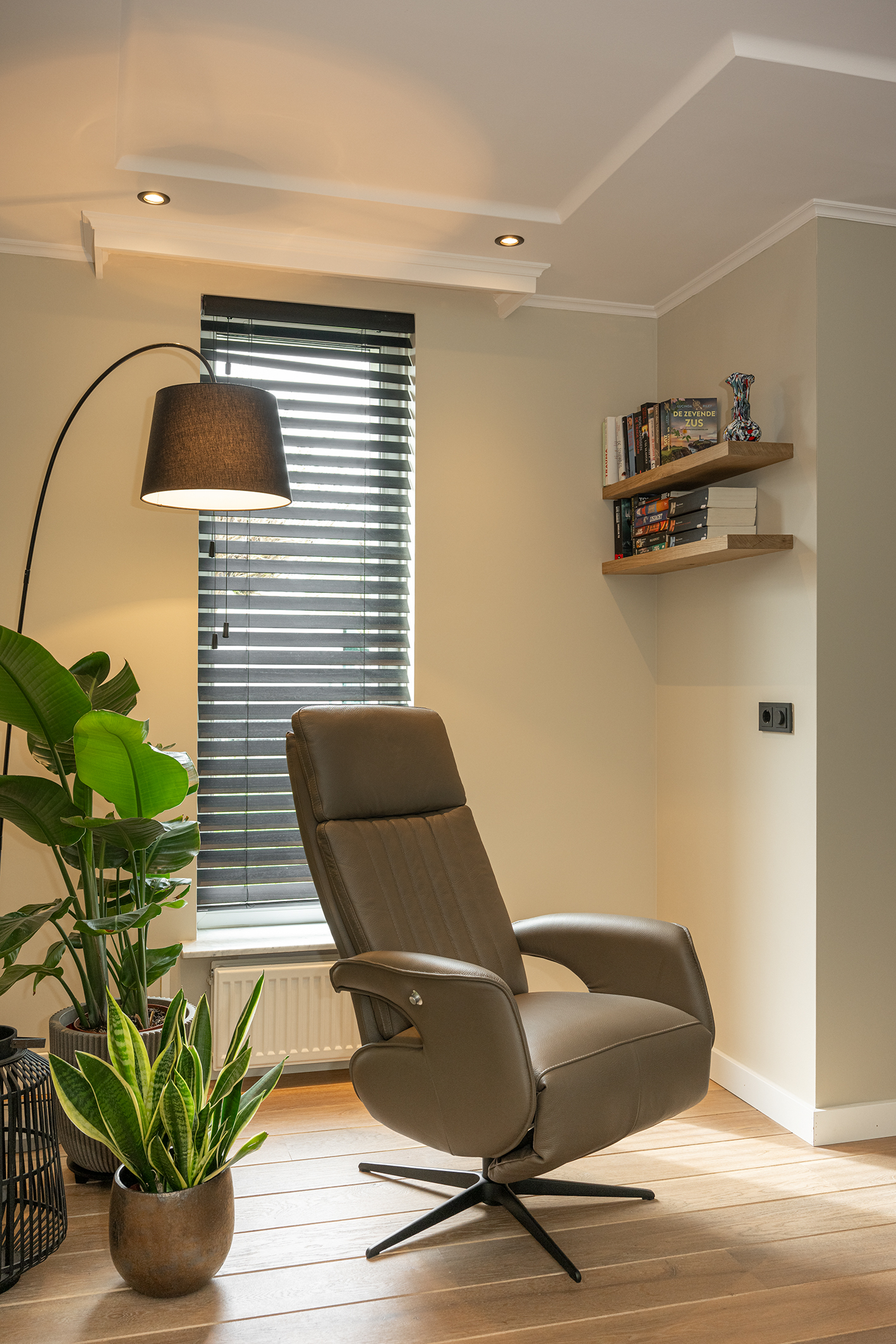Sfeervolle leeshoek met een bruine leren relaxfauteuil, zwarte houten jaloezieën, een booglamp en groene kamerplanten.