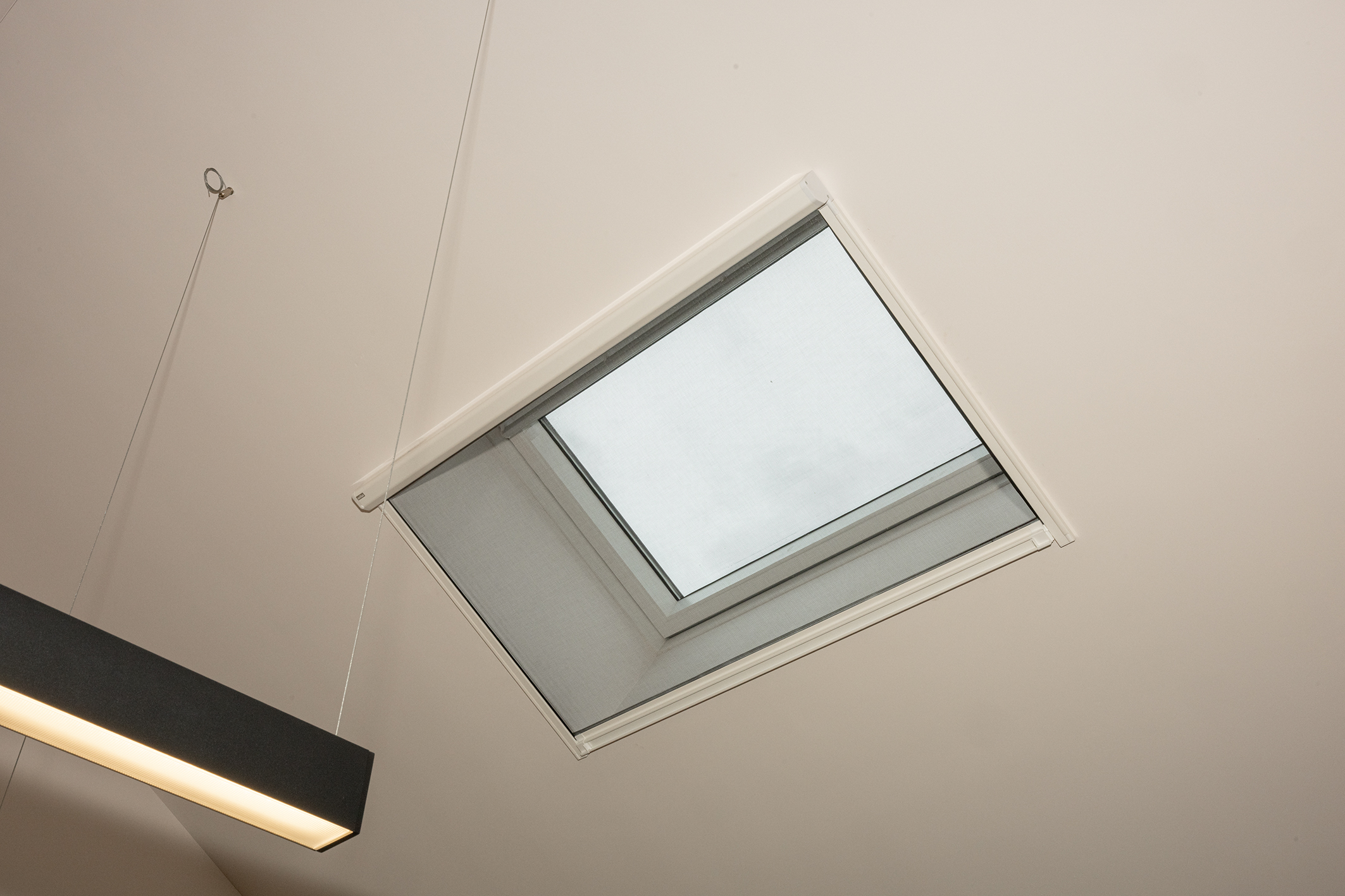 Een vierkant dakraam in een wit plafond, voorzien van een hor en zicht op een bewolkte lucht.