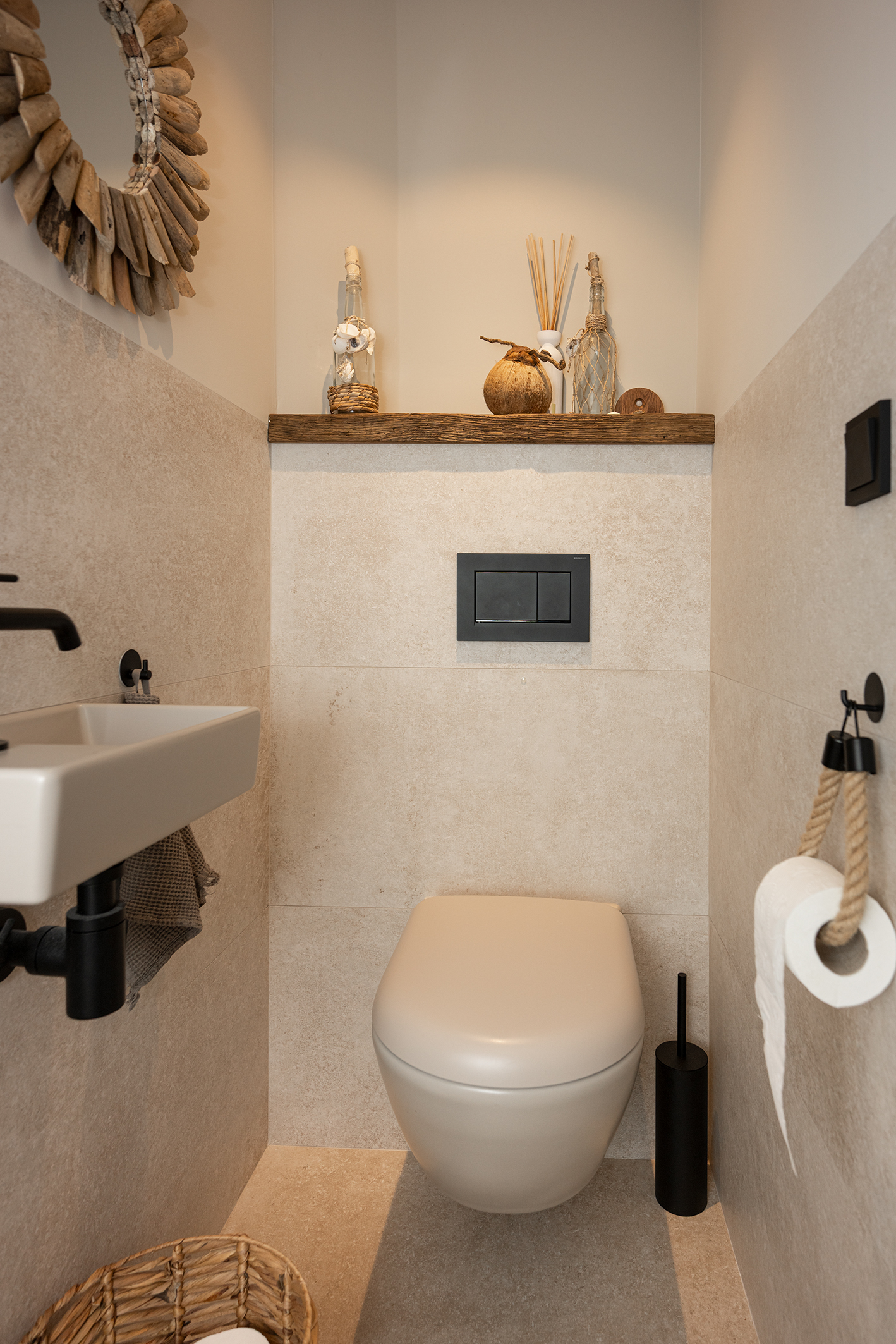 Interieur van een modern toilet met zandkleurige tegels, een zwevend toilet, een houten wandplank met decoratie en een ronde spiegel met een rand van drijfhout.