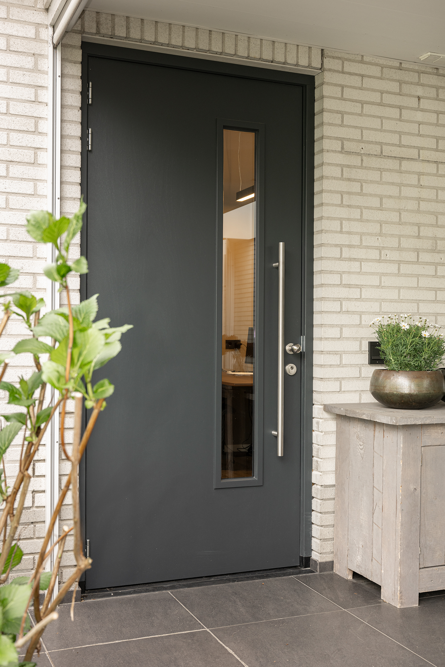 Strakke antracietgrijze voordeur met een verticale glasstrook en een lange RVS handgreep bij een witte bakstenen gevel.