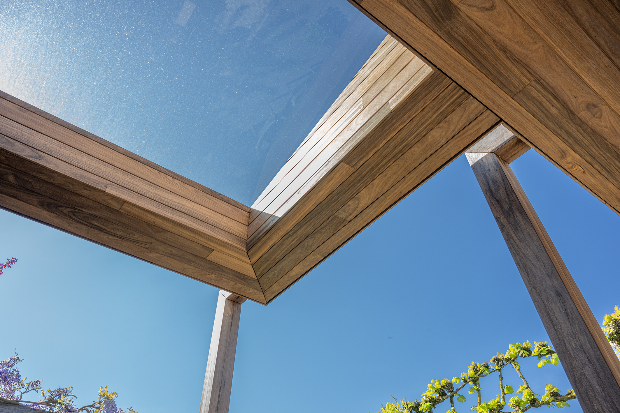 Hoek van de dakconstructie van padoek hout tegen een strakblauwe lucht met glazen dakvenster.