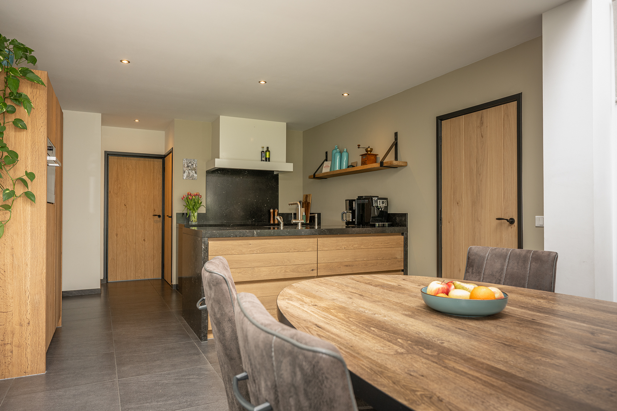 Overzicht van een moderne open keuken met een houten eettafel, een kookeiland met donker blad en houten deuren met zwarte kozijnen.