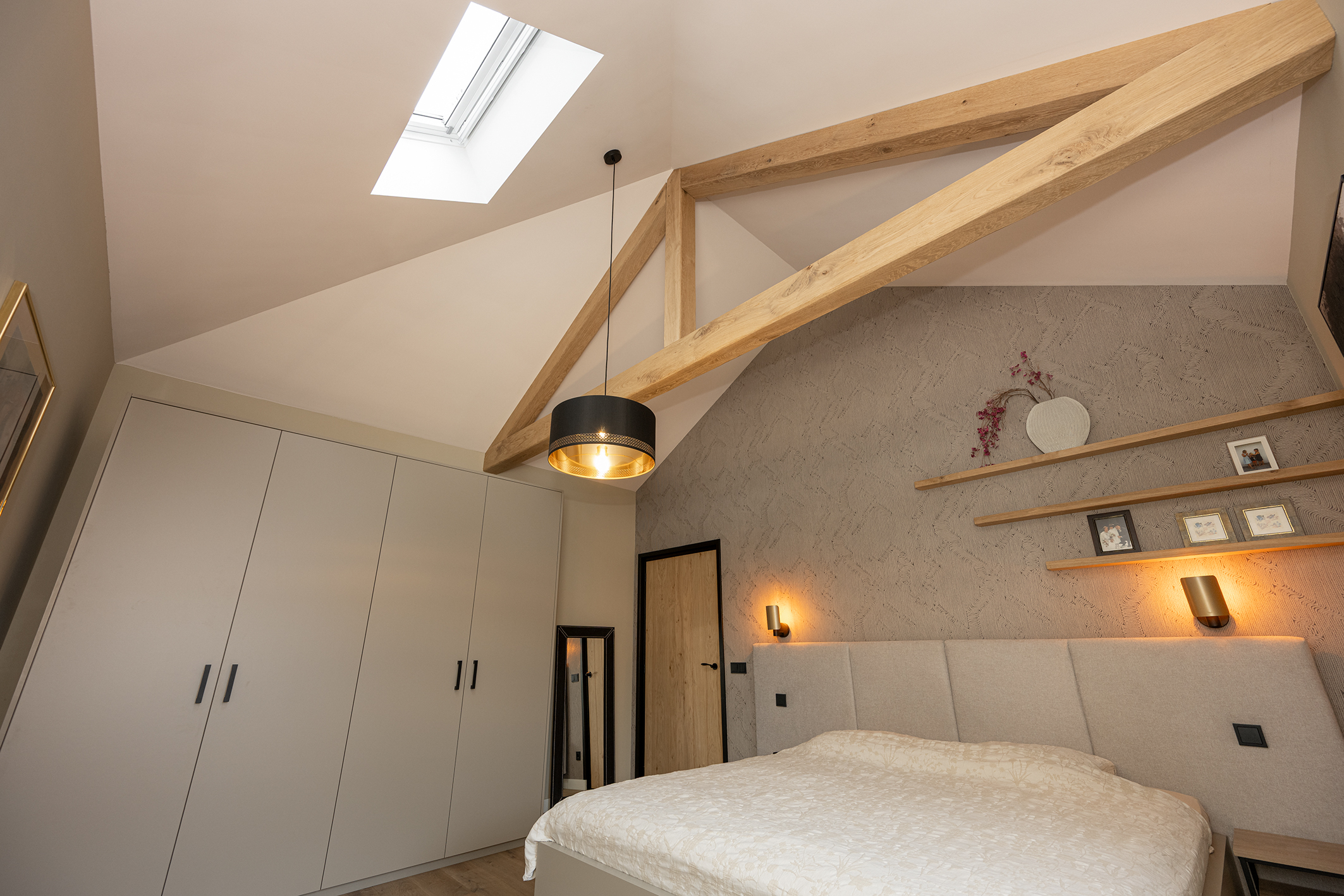 Overzicht van een moderne slaapkamer met een hoog schuin plafond, robuuste houten steunbalken, een dakraam en een grote grijze kledingkast.