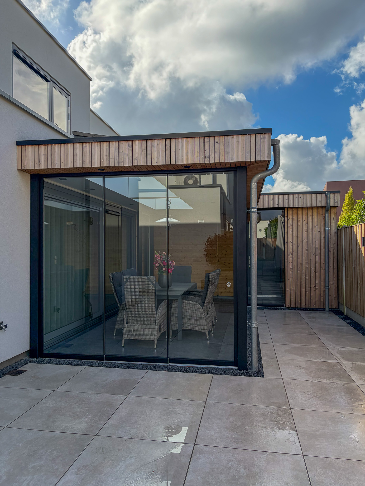 Een breed perspectief van een moderne tuinoverkapping met gesloten glazen schuifwanden en een betegeld terras onder een bewolkte hemel.