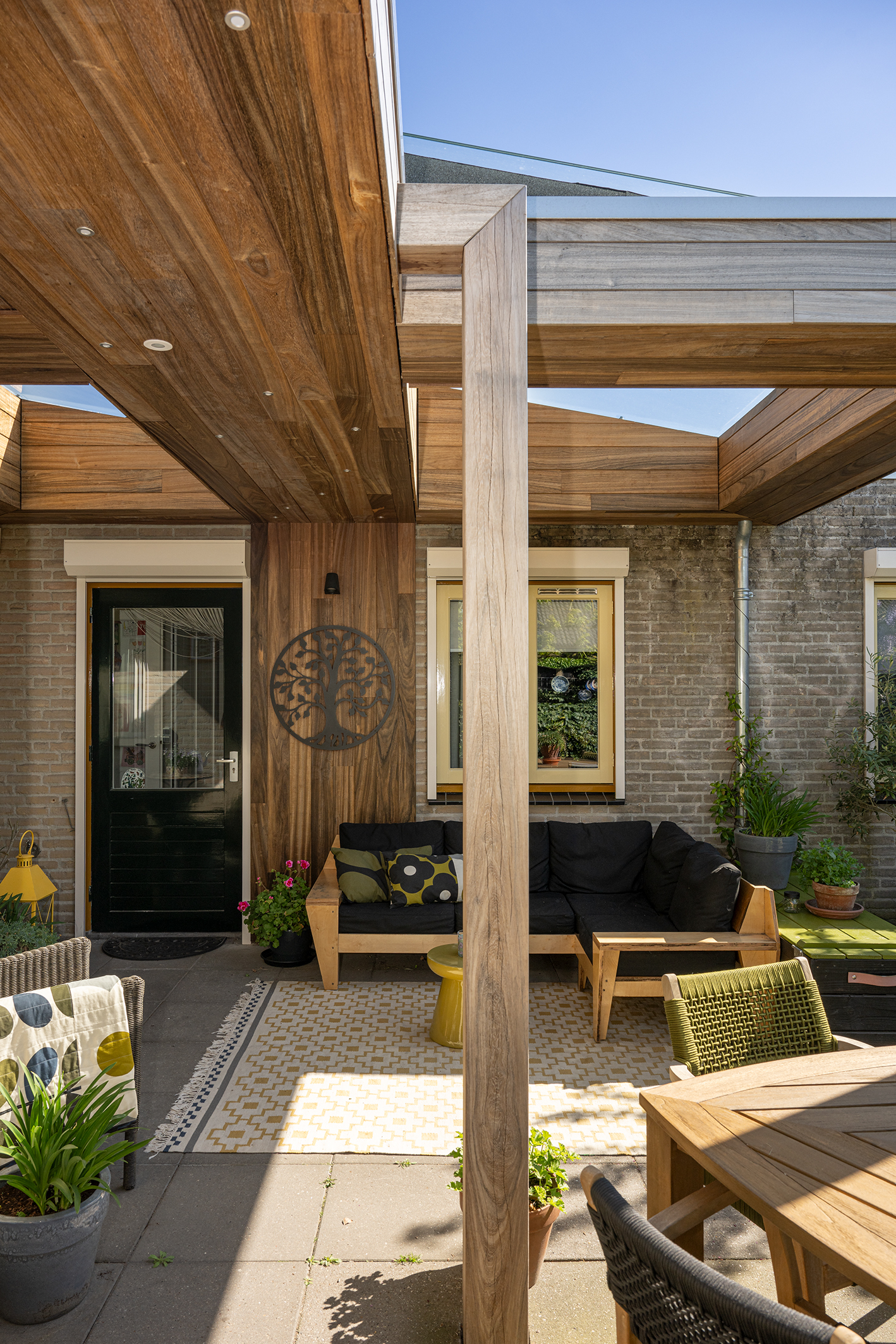 Wand van padoek hout met een metalen levensboom kunstwerk onder een terrasoverkapping.