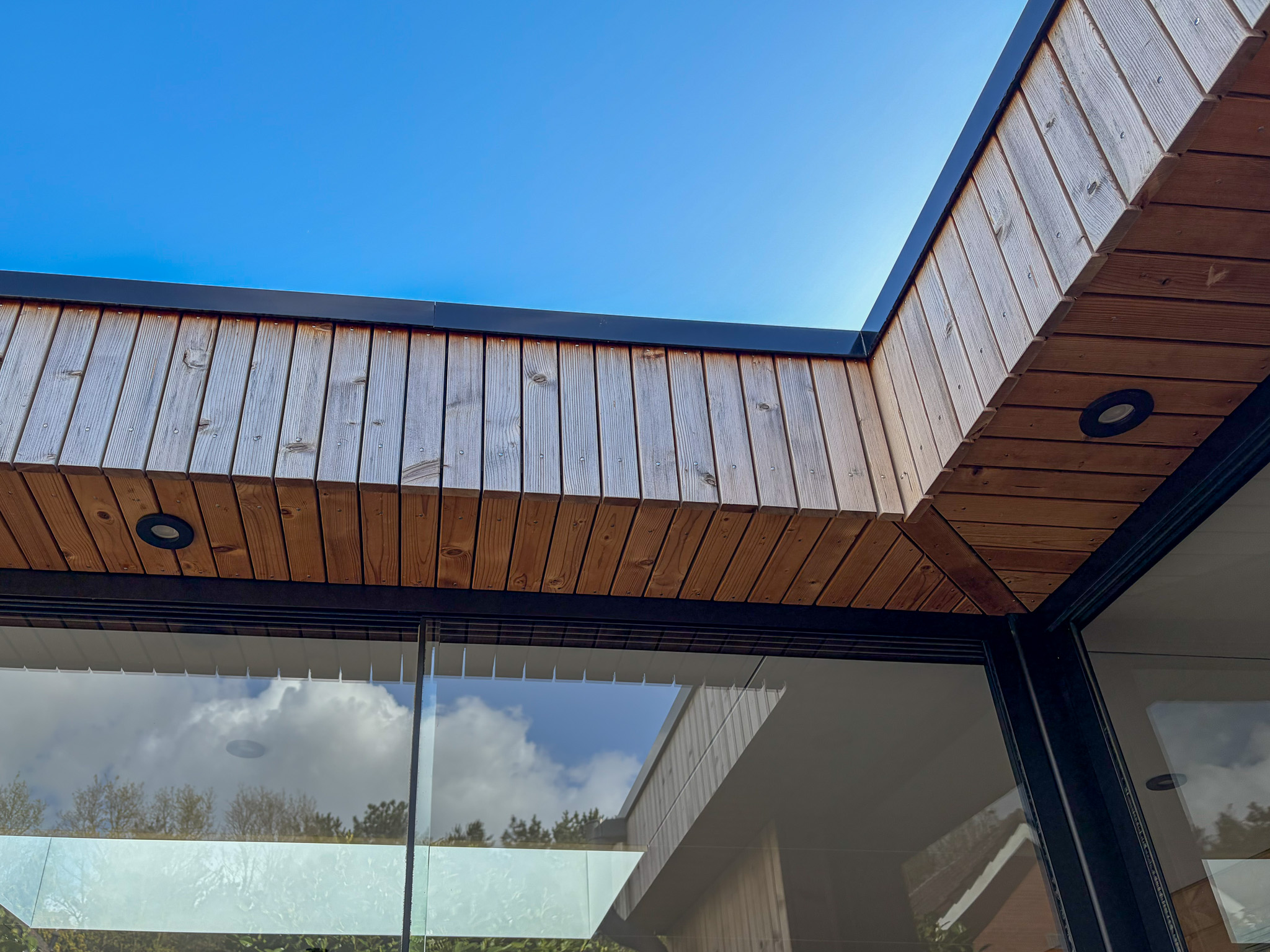 Onderkant van het houten plafond van de overkapping met ingebouwde zwarte spotjes tegen een blauwe lucht.